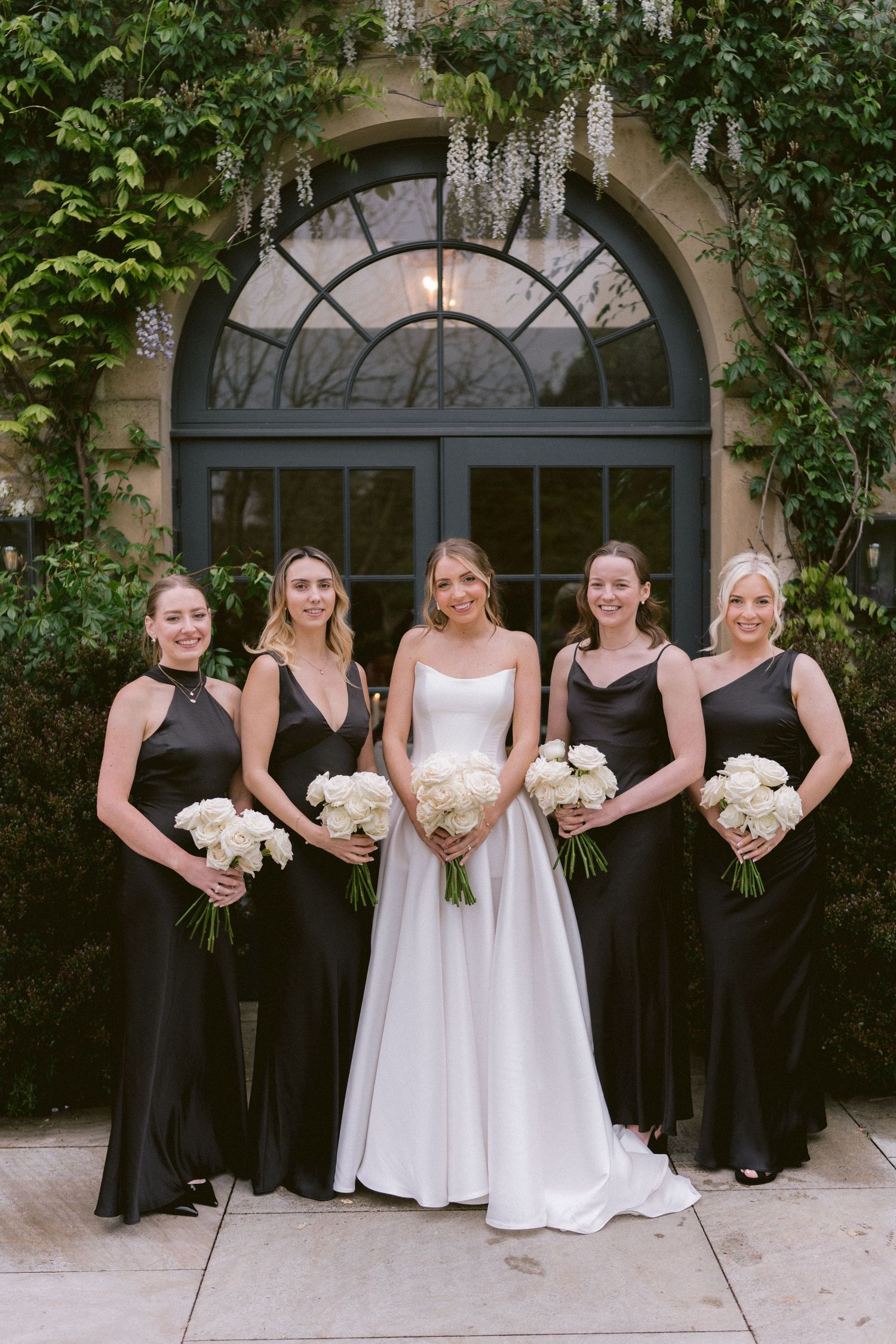 Bride in white gown poses with four bridesmaids in black dresses holding white bouquets before an ivy-covered building.