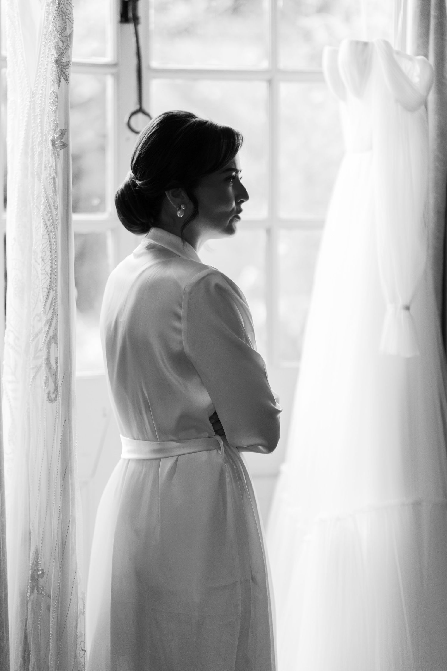 Bride in white robe gazes at her wedding dress hanging by a window in a black and white photo.