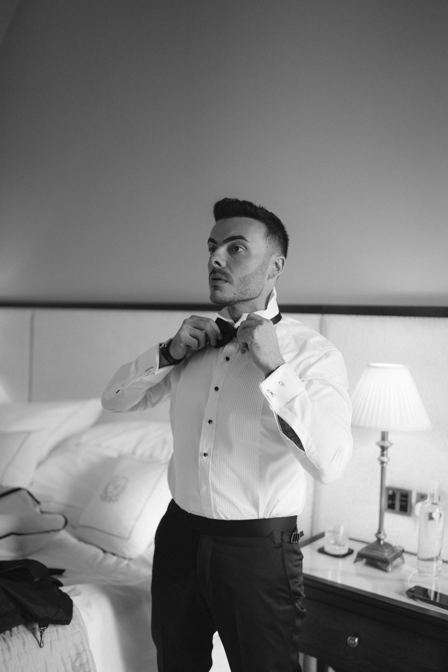 Groom in white dress shirt adjusting bow tie in hotel room, black and white photo.
