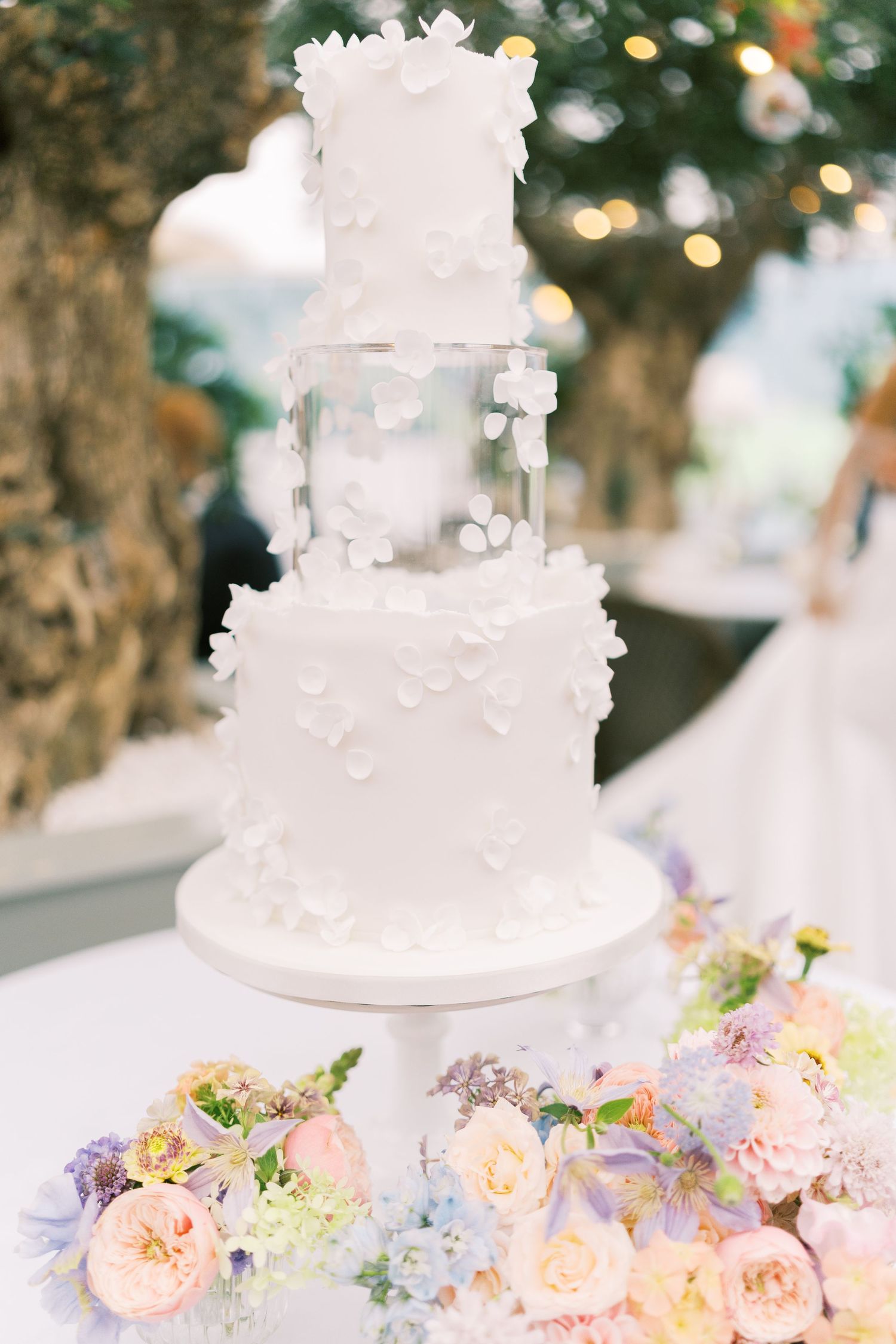 Four-tier white wedding cake adorned with floral appliques surrounded by pastel roses and blooms.