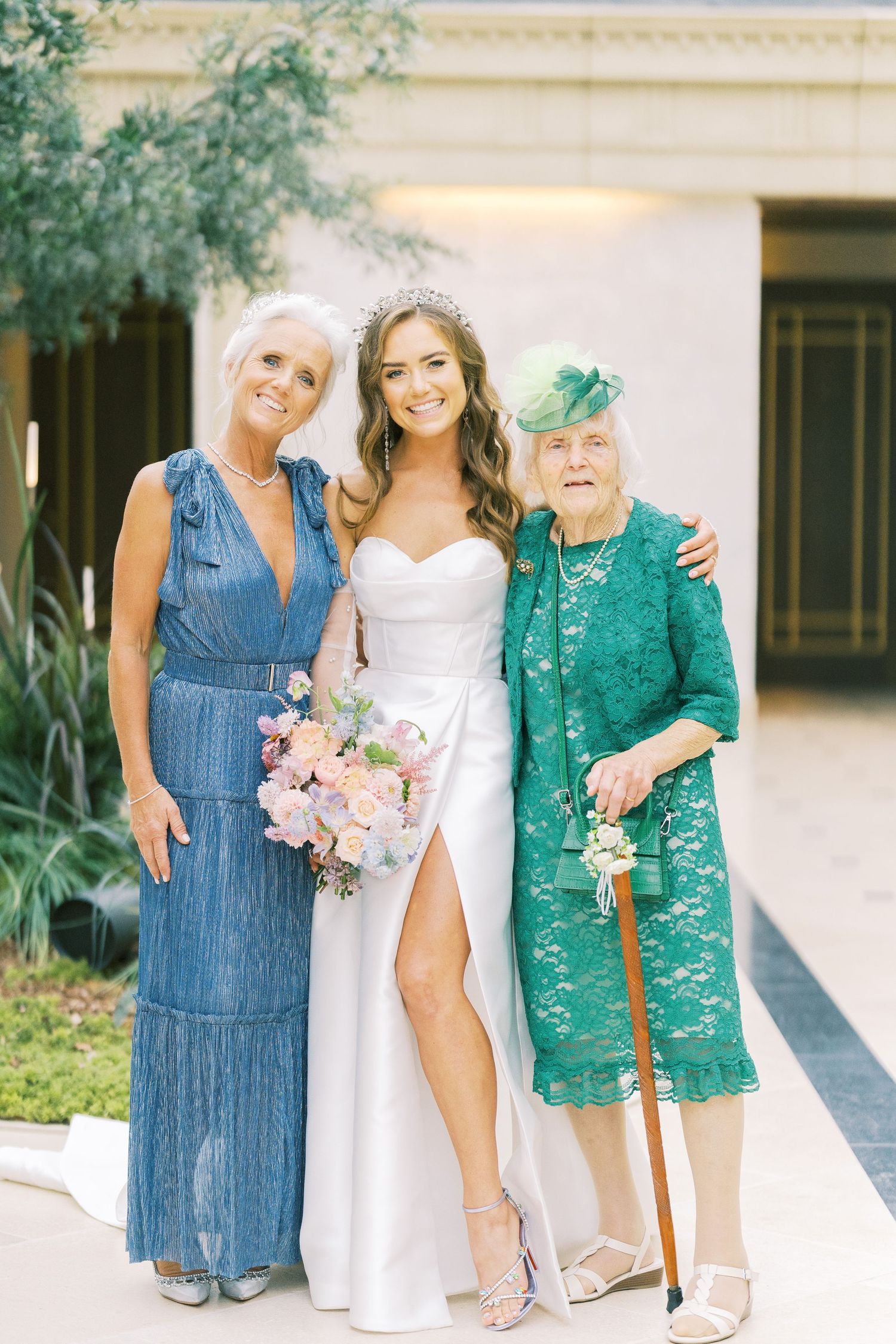 Bride in white gown with bouquet poses with mother in blue dress and grandmother in green lace at wedding.