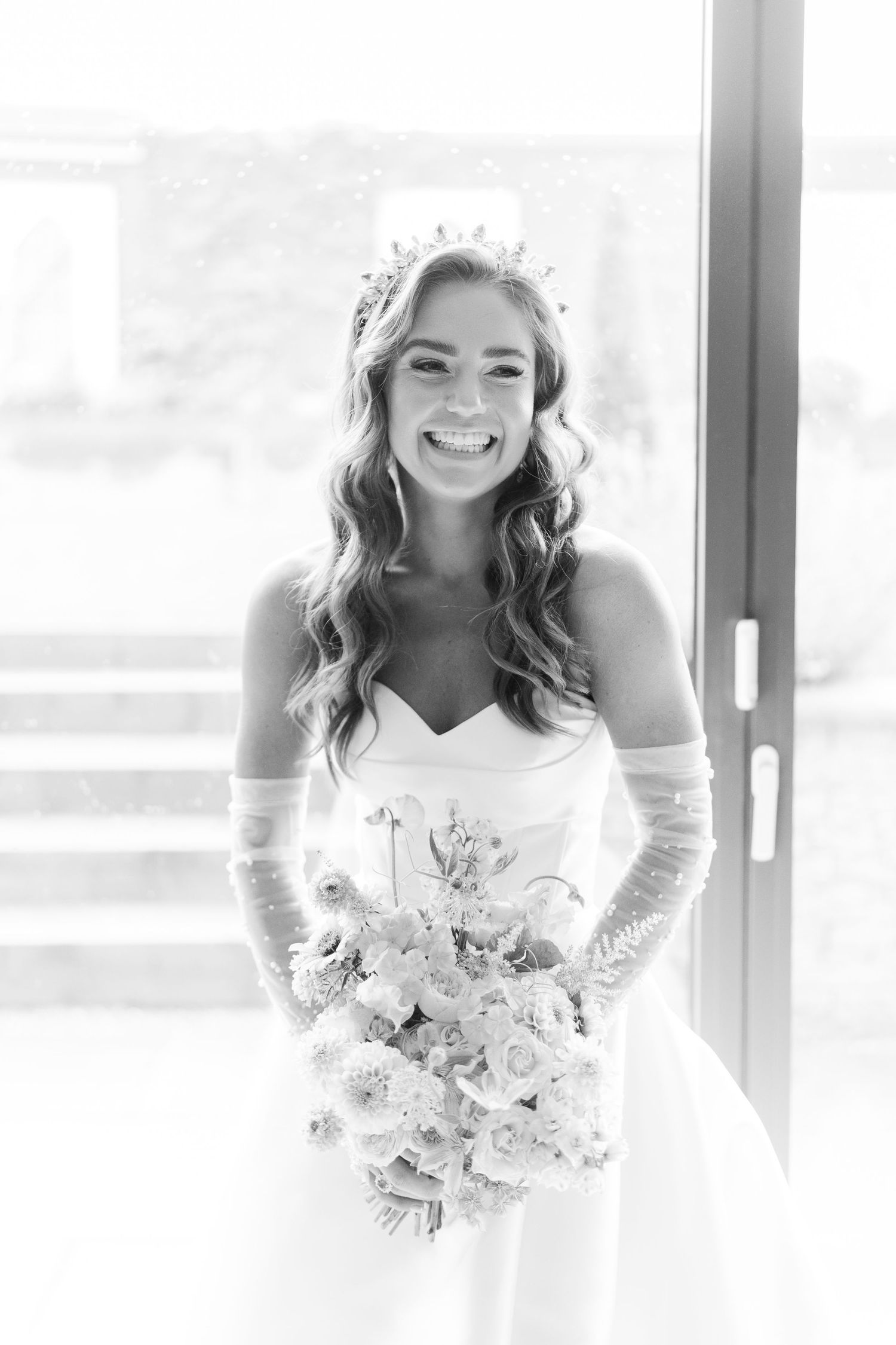 Smiling bride in strapless gown with tiara and gloves holding a bouquet in black and white photo.