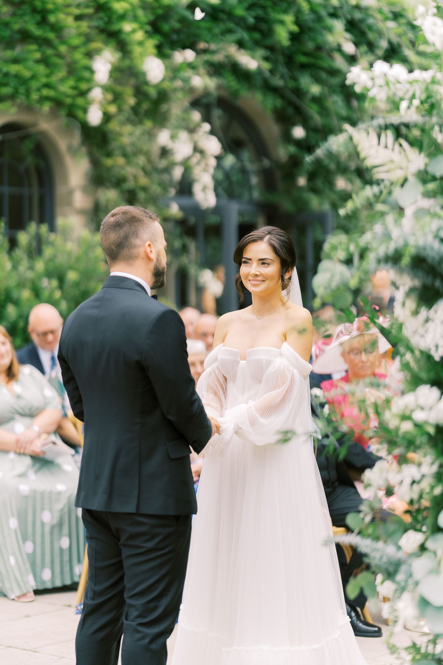 Bride in off-shoulder white gown smiling at groom during outdoor wedding ceremony surrounded by lush greenery.