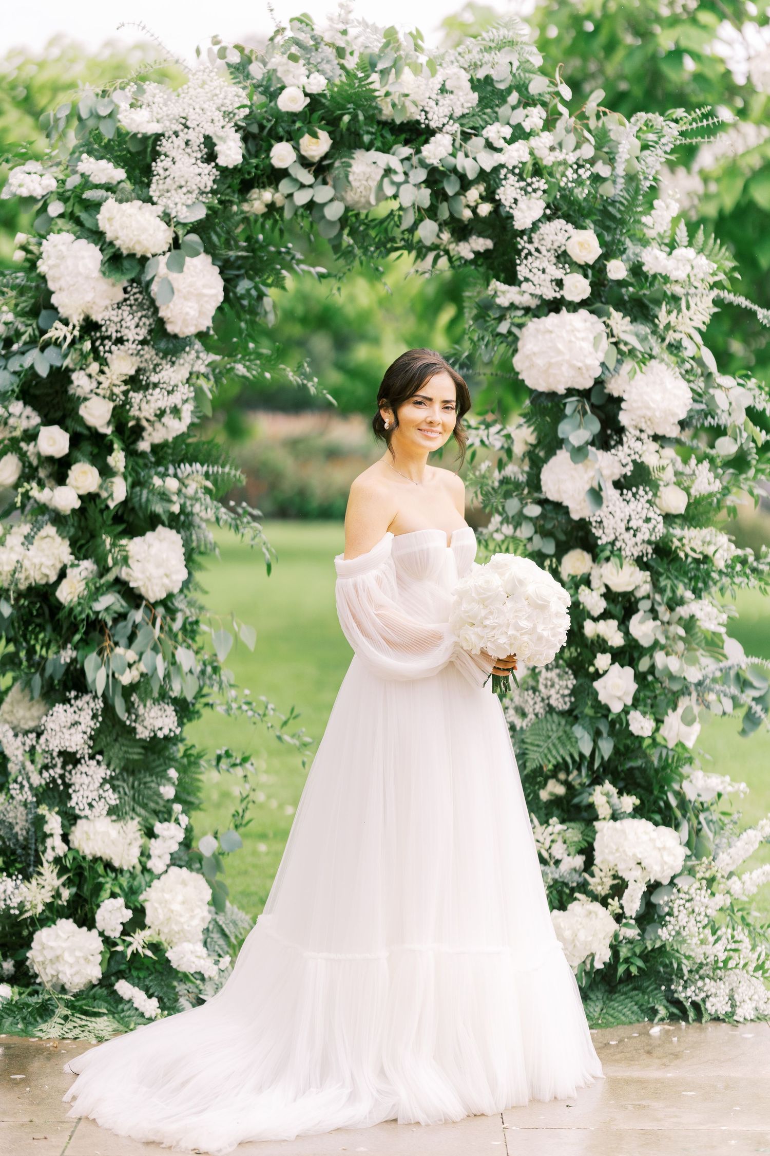 Smiling bride in off-shoulder gown holds white bouquet under floral arch with greenery.