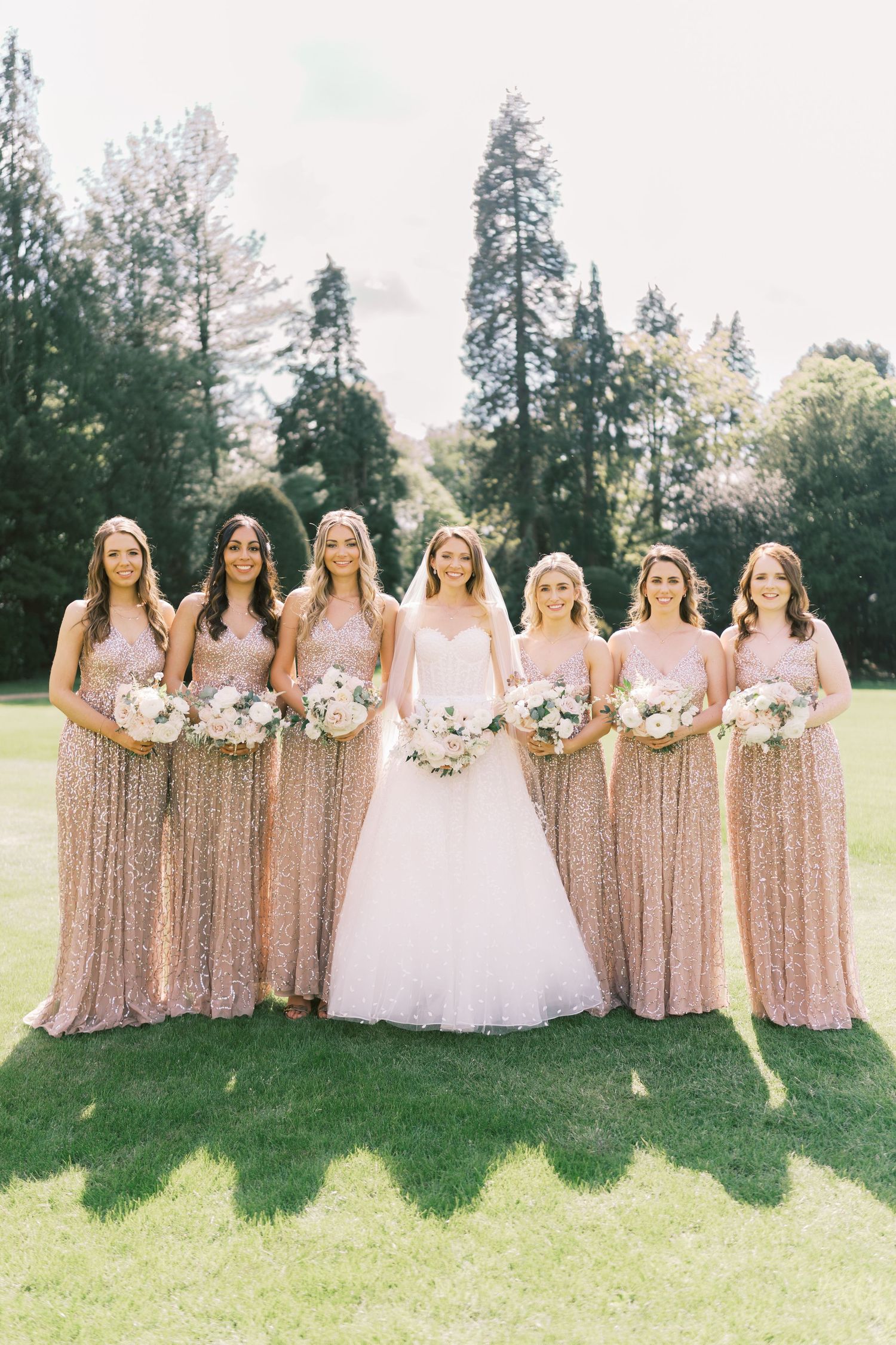 Bride in white ballgown poses with six bridesmaids in rose gold sequin dresses holding white bouquets outdoors.