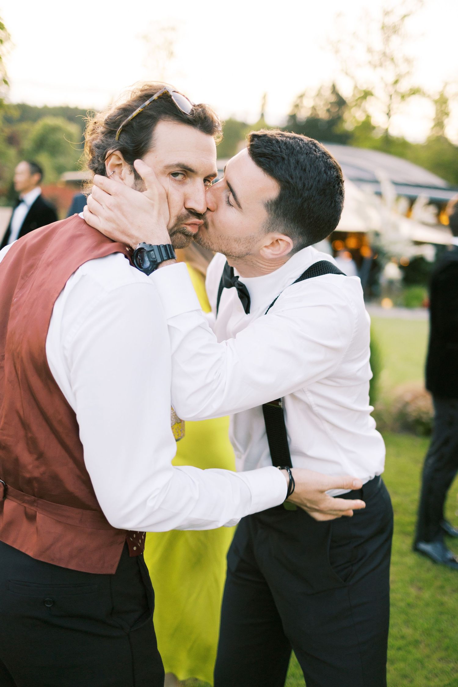 Two grooms share a kiss at an outdoor wedding reception, one wearing a burgundy vest and the other a black bow tie.