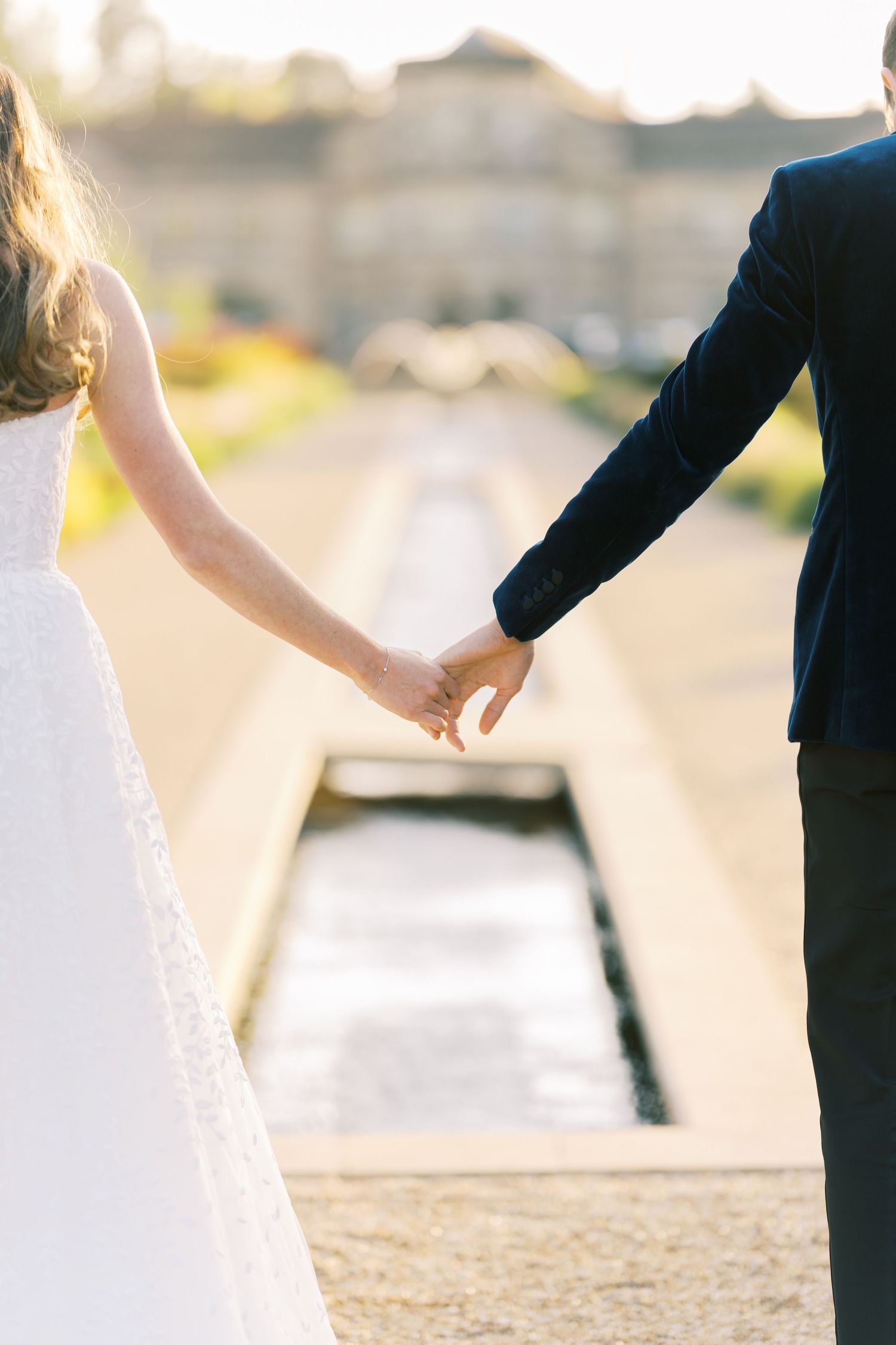 Close-up of bride and groom holding hands near a garden water channel at a golden hour wedding.
