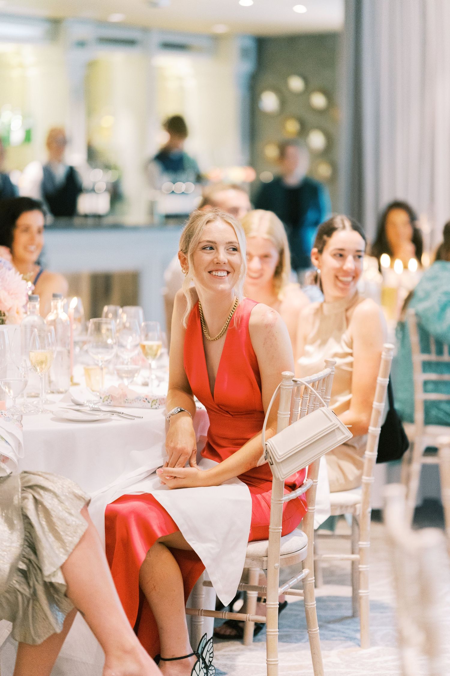 Smiling woman in red dress seated at elegant wedding reception table with guests laughing in background.