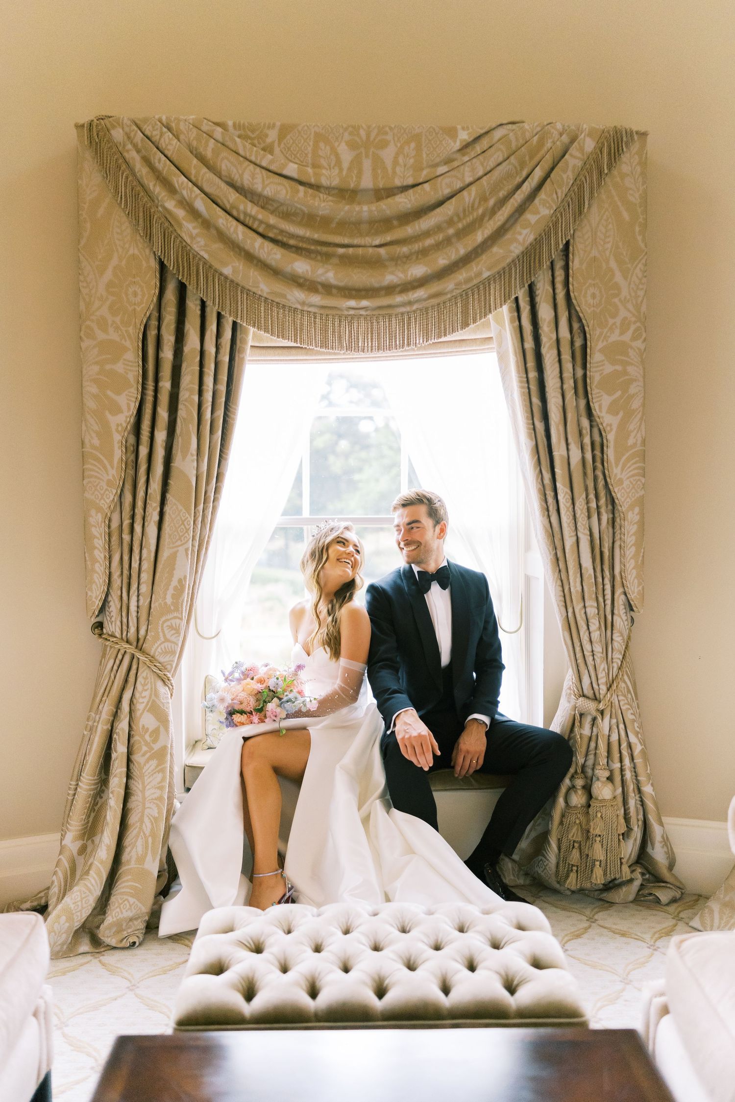 Elegant bride and groom sitting together in window seat framed by ornate gold damask curtains on wedding day.