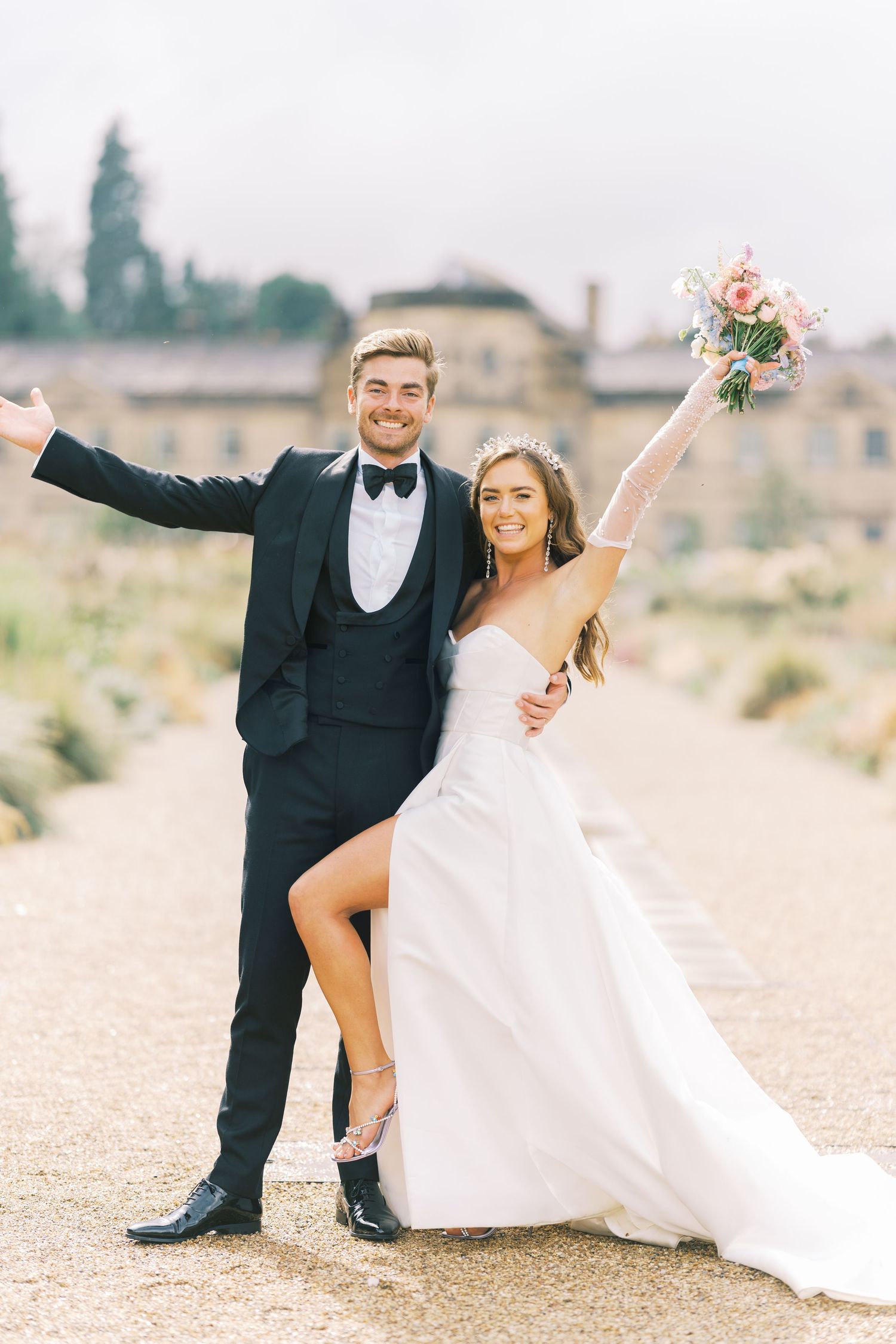 Happy bride and groom celebrating outdoors, bride raising bouquet in white gown and tiara, groom in black tuxedo.