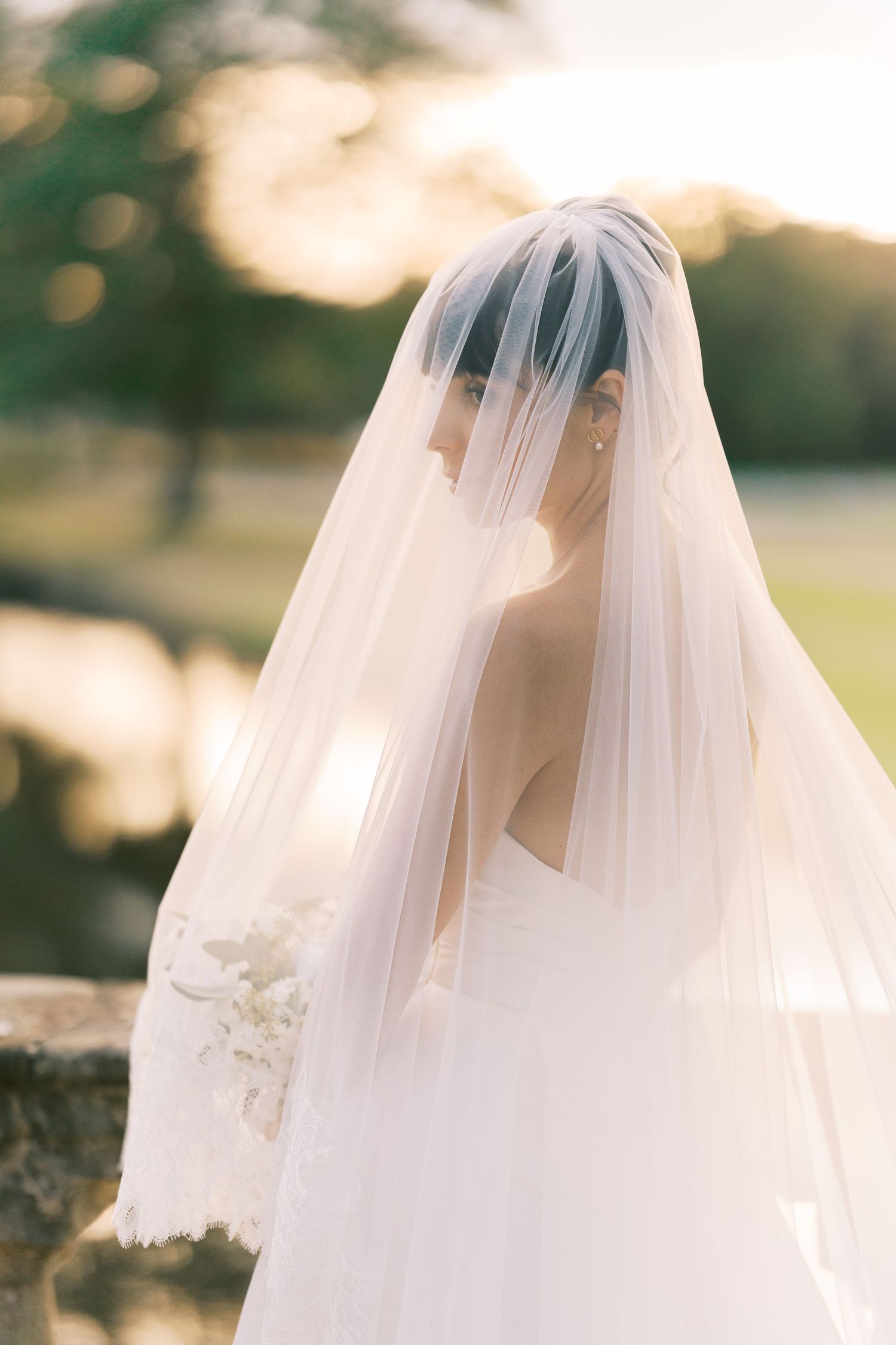 Bride in a white lace gown and flowing veil poses outdoors at golden hour, holding a white bouquet.