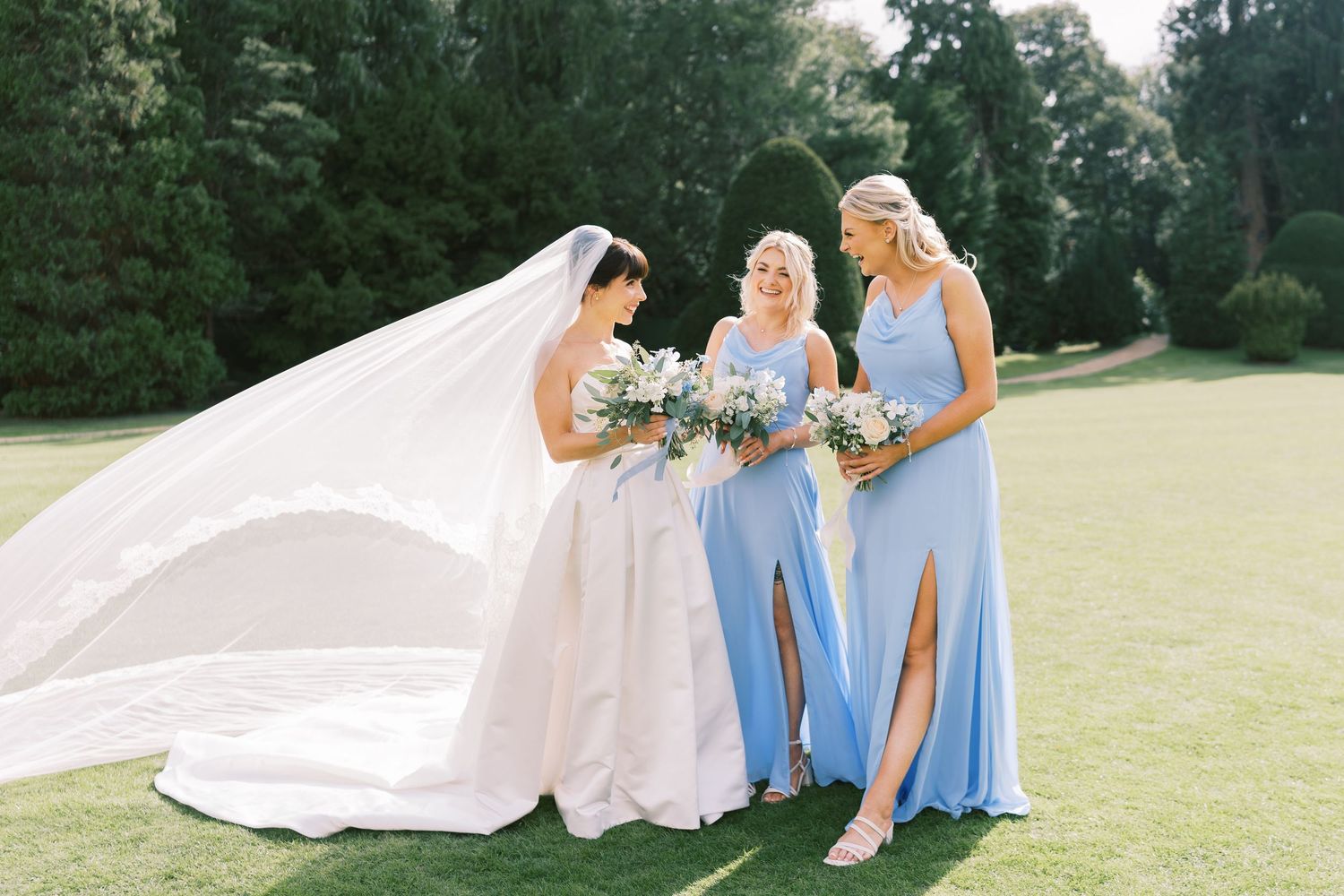 Bride in white gown with flowing veil stands with two bridesmaids in blue dresses holding bouquets on green lawn.