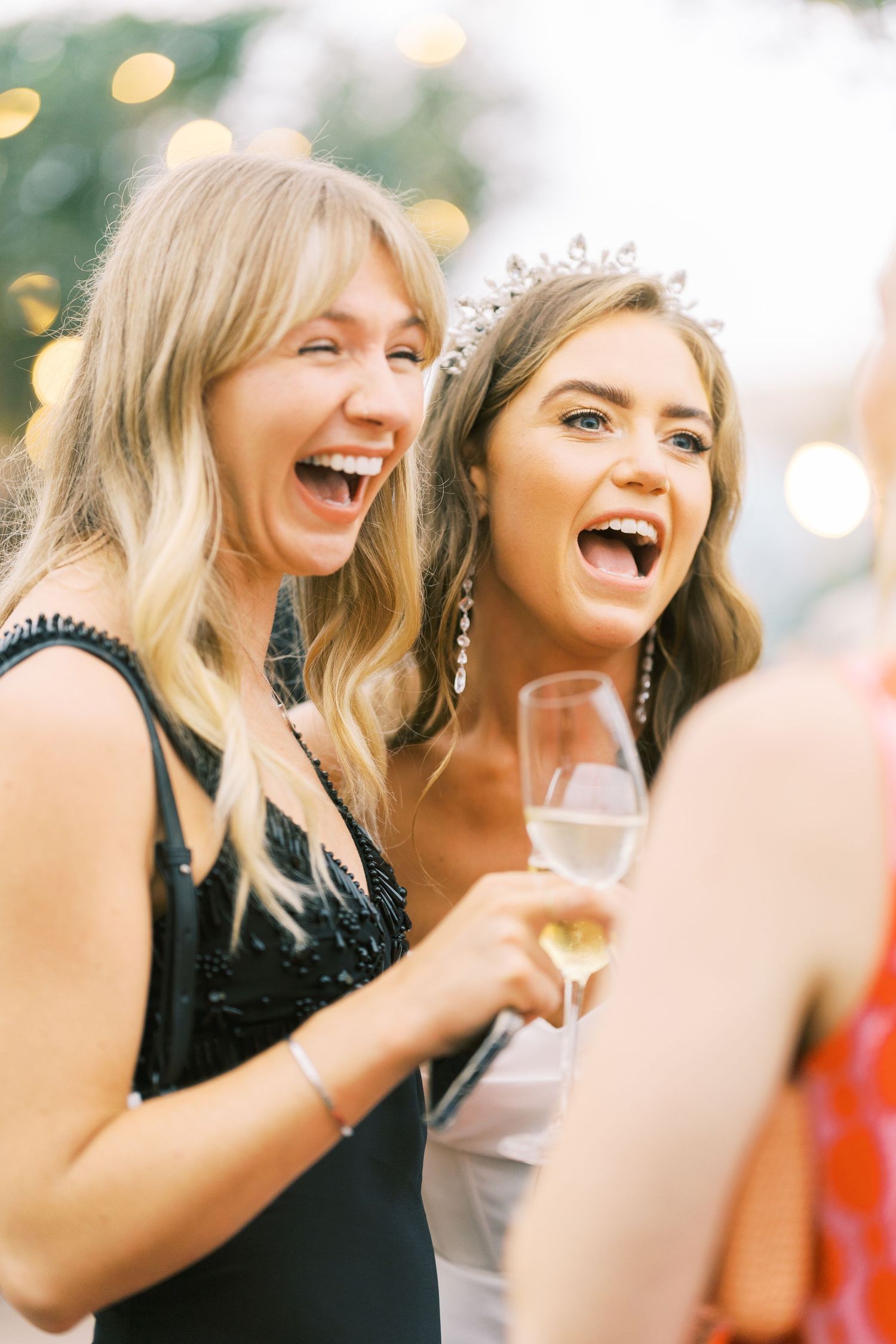 Laughing bride wearing diamond tiara clinks champagne glasses with blonde guest at outdoor wedding reception.