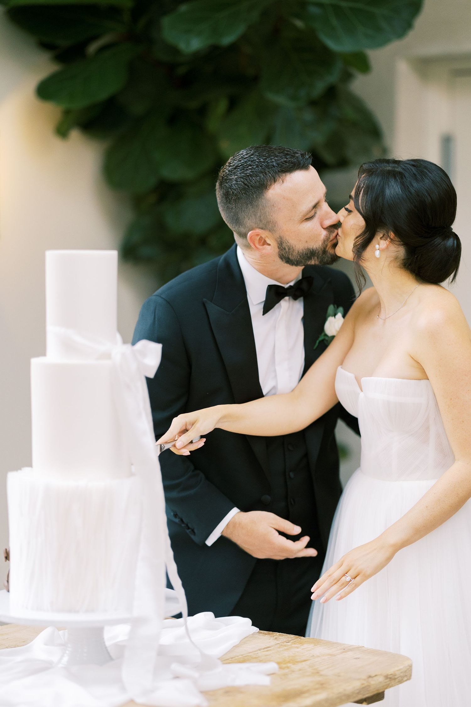 Bride and groom sharing a kiss while cutting their elegant white tiered wedding cake together.