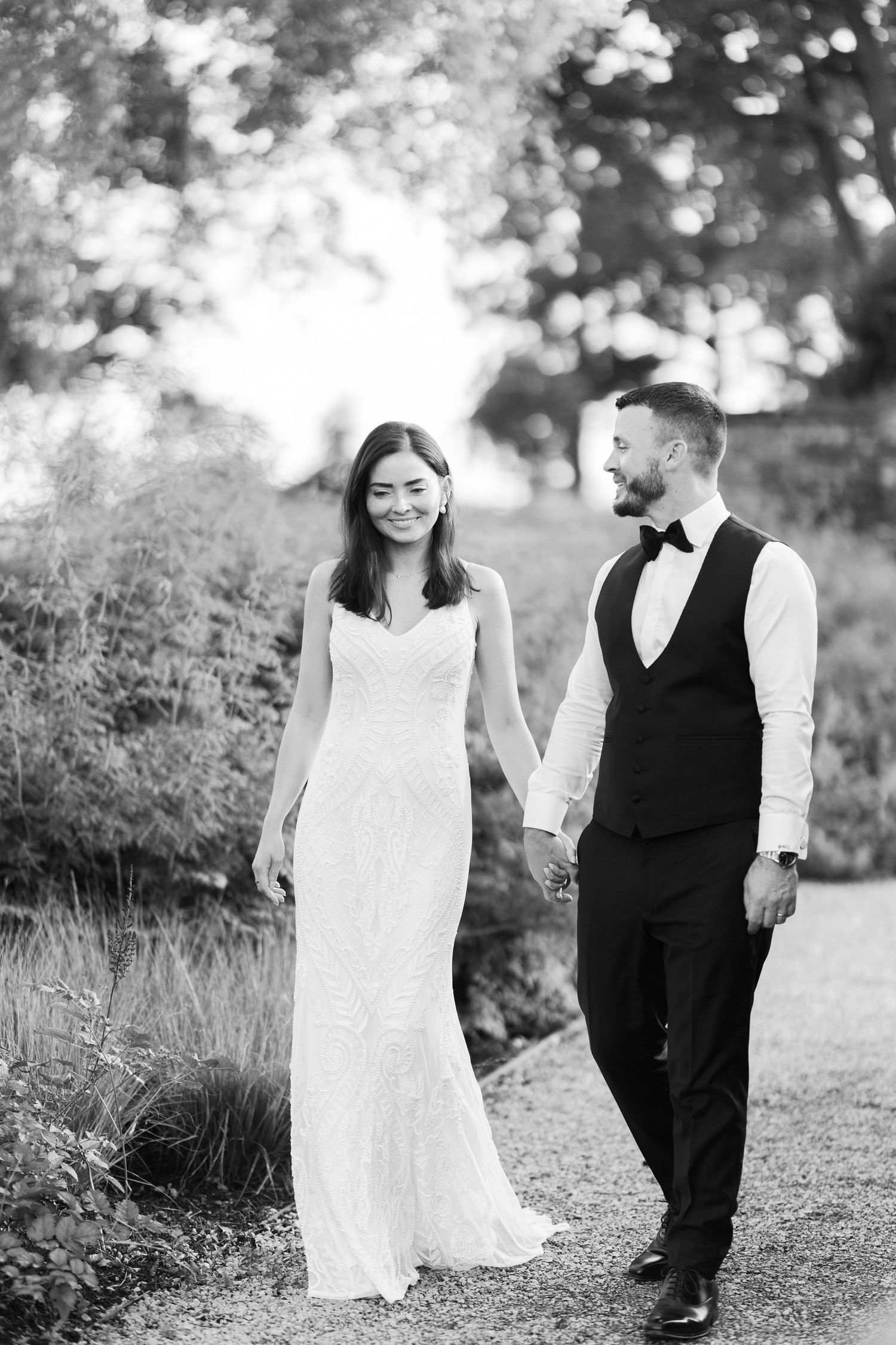 Black and white photo of a smiling bride and groom holding hands while walking along a garden path.