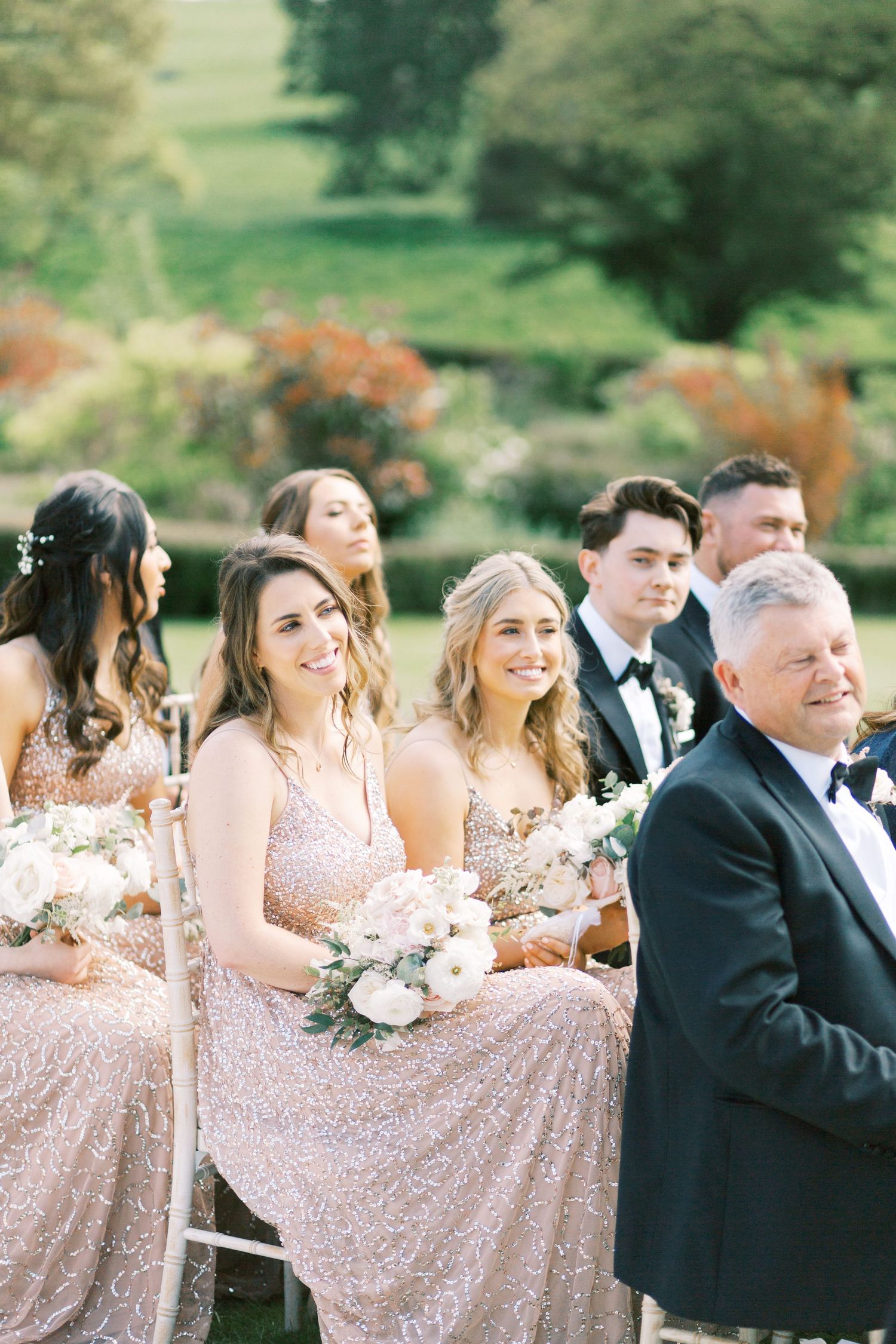 Smiling bridesmaids in rose gold sequin dresses holding white bouquets seated at an elegant outdoor wedding ceremony.