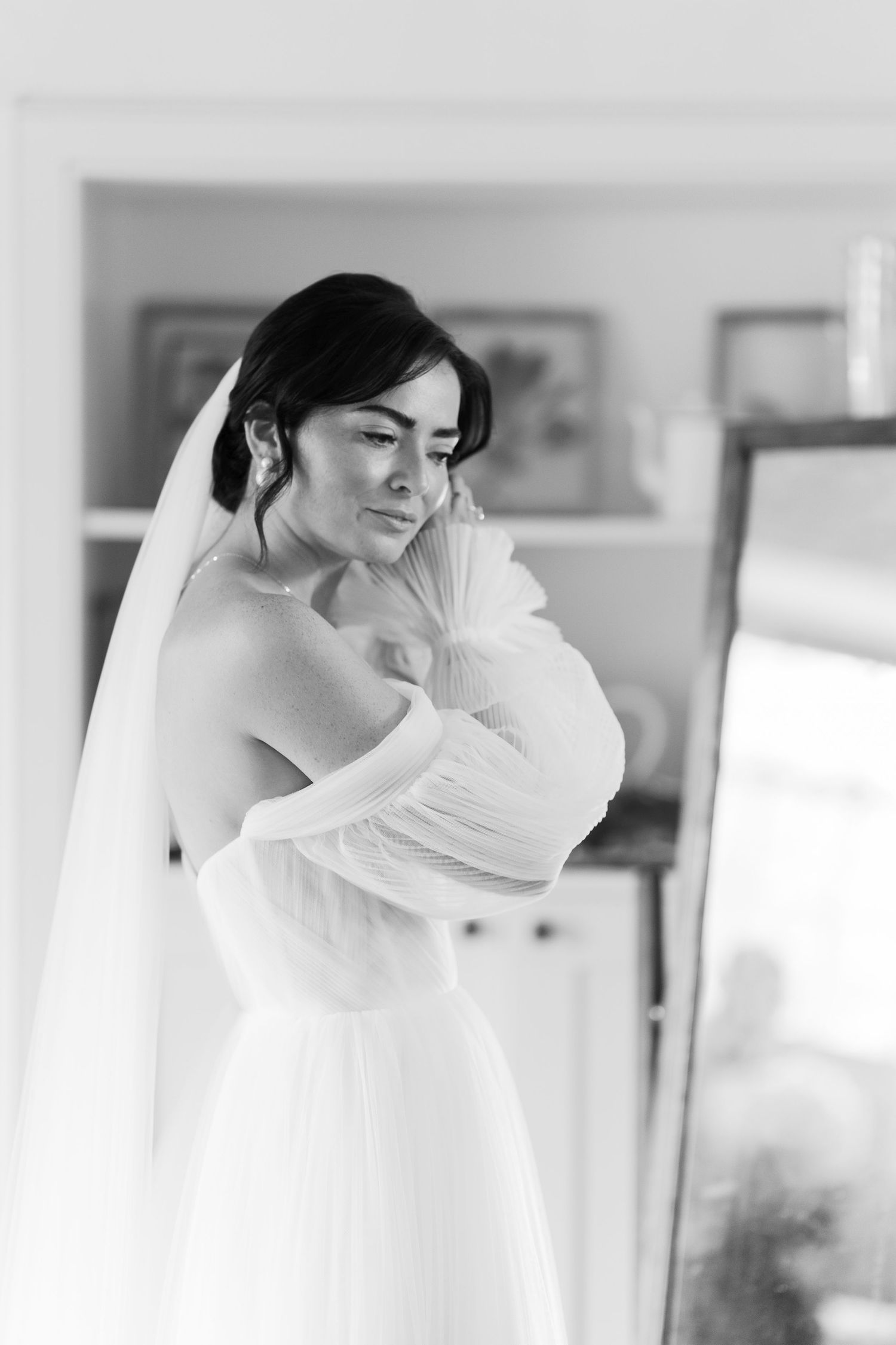 Black and white portrait of bride in off-shoulder wedding gown and veil posing near mirror.