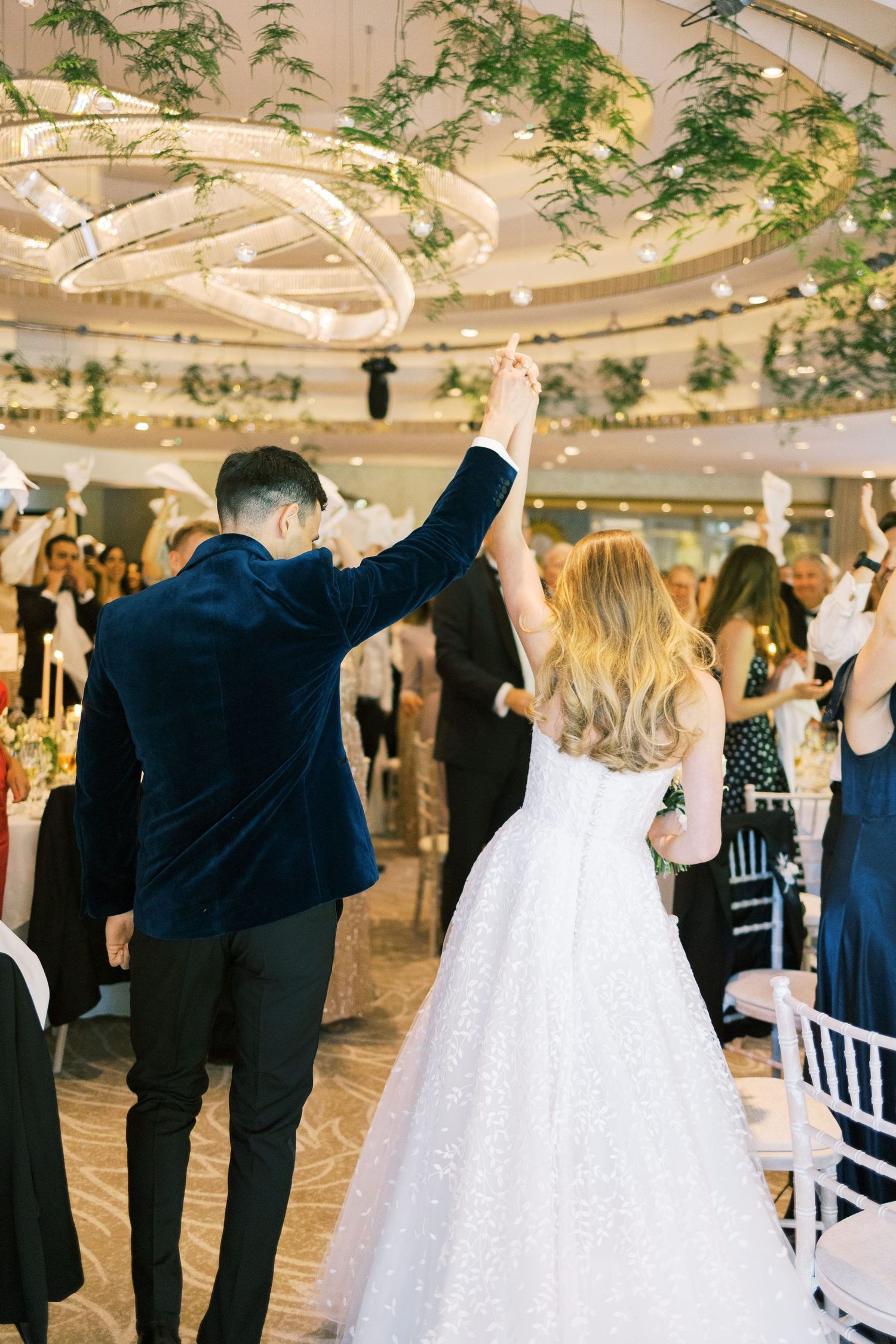Bride and groom raise hands entering wedding reception hall with elegant chandelier and greenery decor.