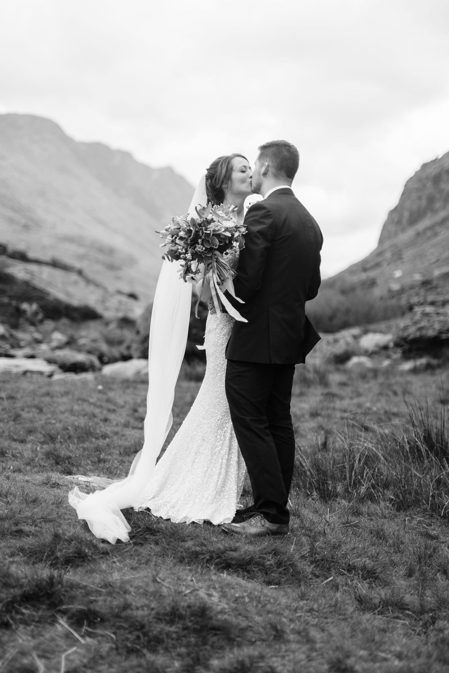 Black and white wedding photo of bride and groom kissing in a scenic mountain landscape.