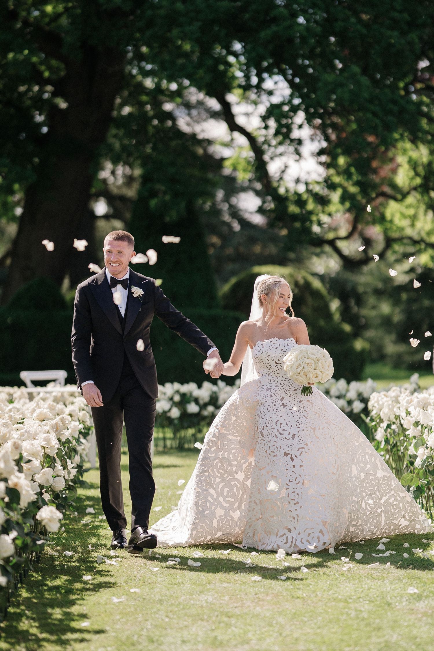 Bride and groom walk hand-in-hand down a flower-lined aisle in a lush garden, bride wearing an ornate ballgown.