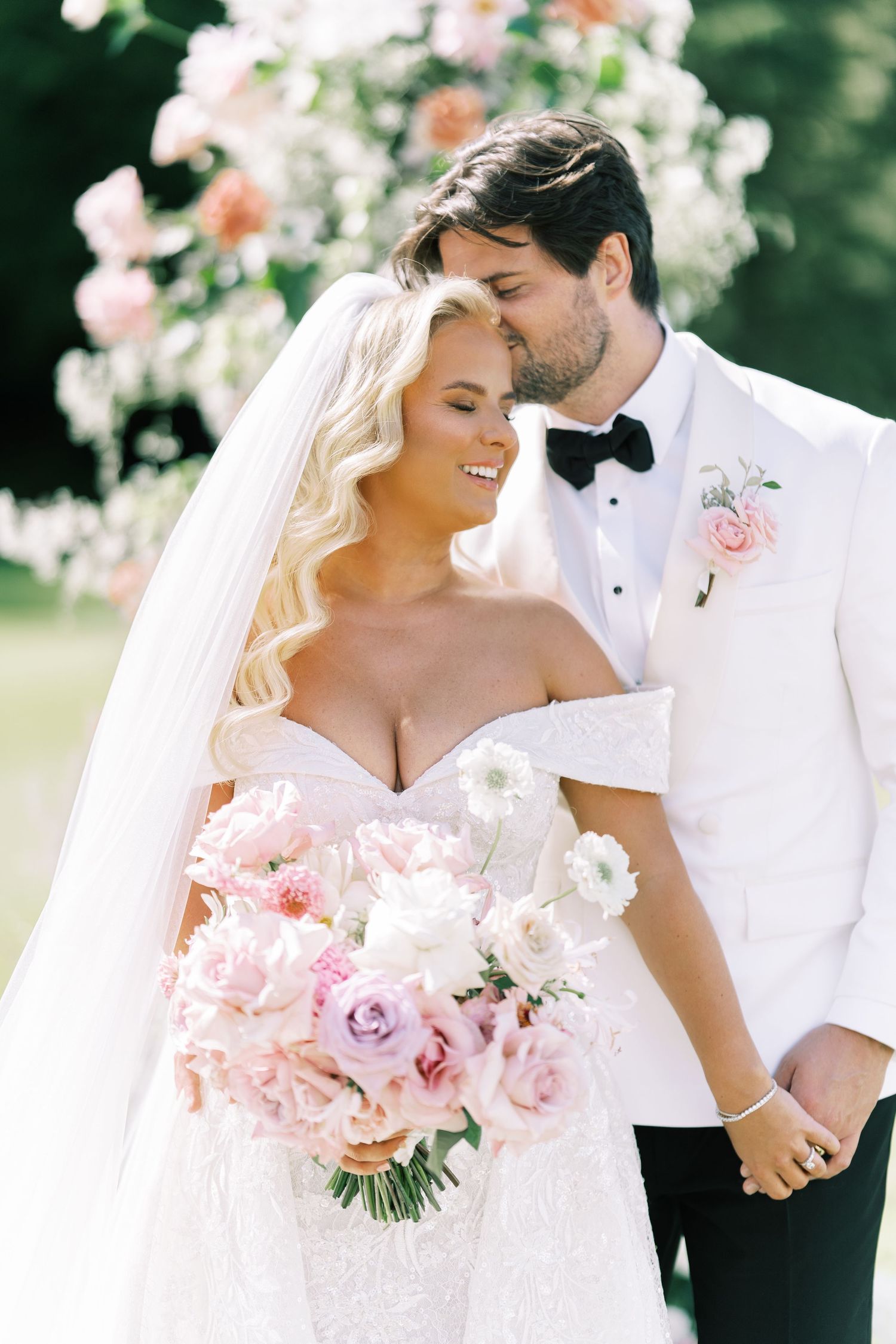 Bride in off-shoulder gown holds pink bouquet as groom in white tuxedo kisses her head at outdoor wedding.