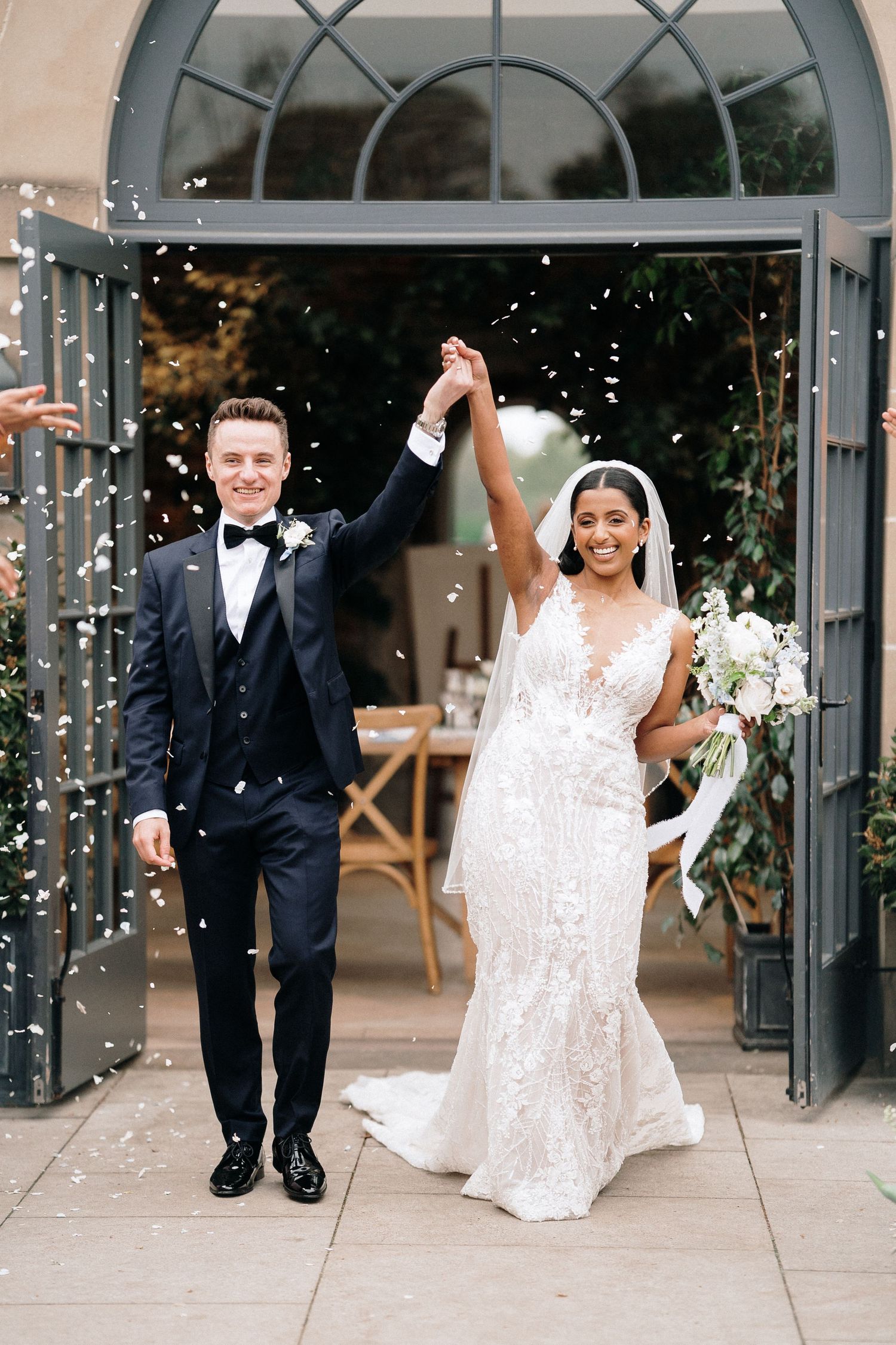 Joyful bride and groom raising hands as confetti falls outside elegant venue doors.