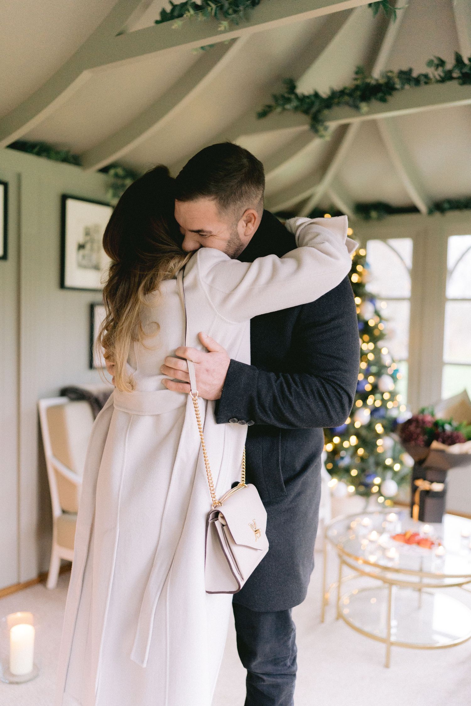 Couple sharing a heartfelt embrace inside a cozy Christmas-decorated room with a glowing tree in the background.