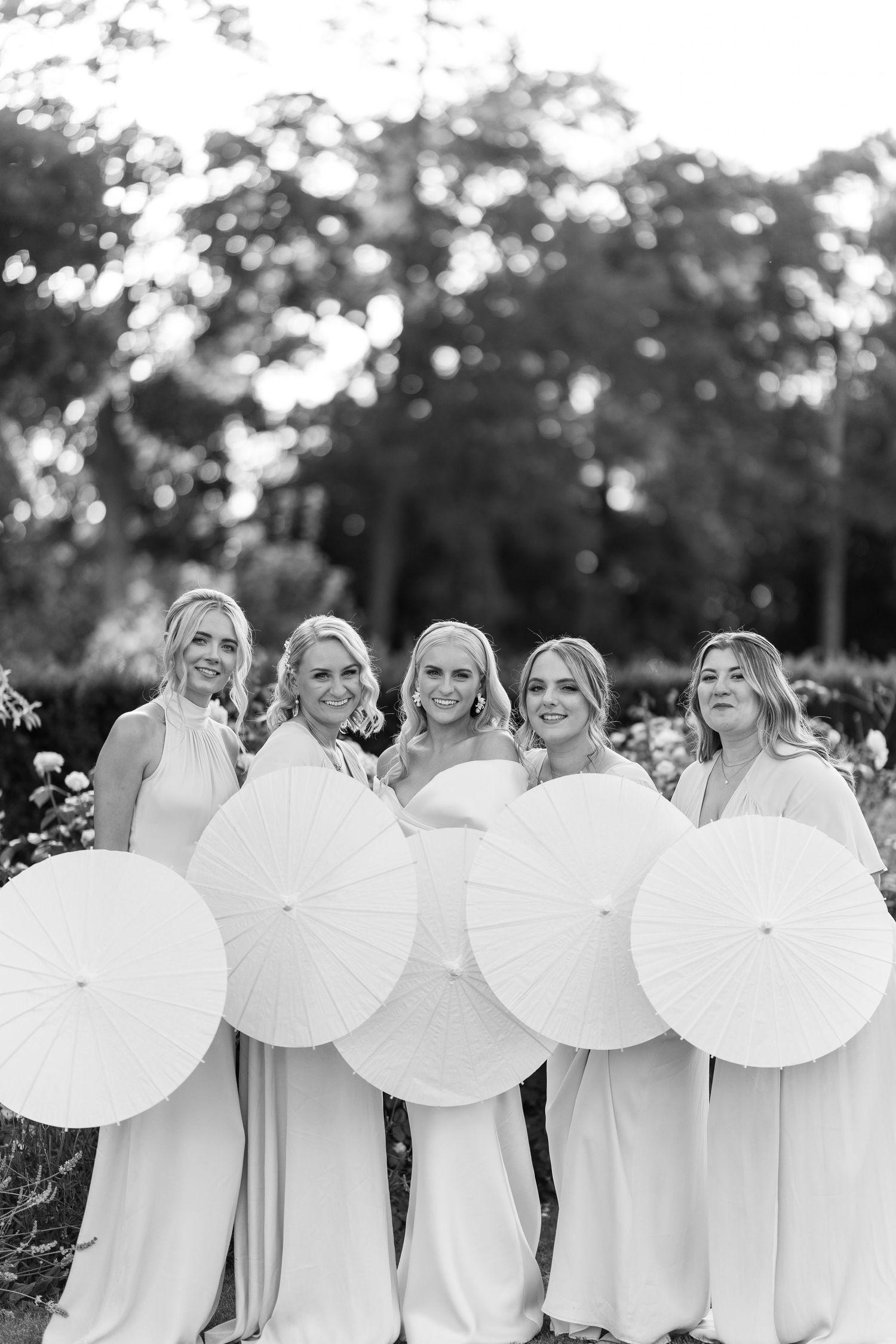 Black and white photo of bride and bridesmaids holding white parasols outdoors in a garden.