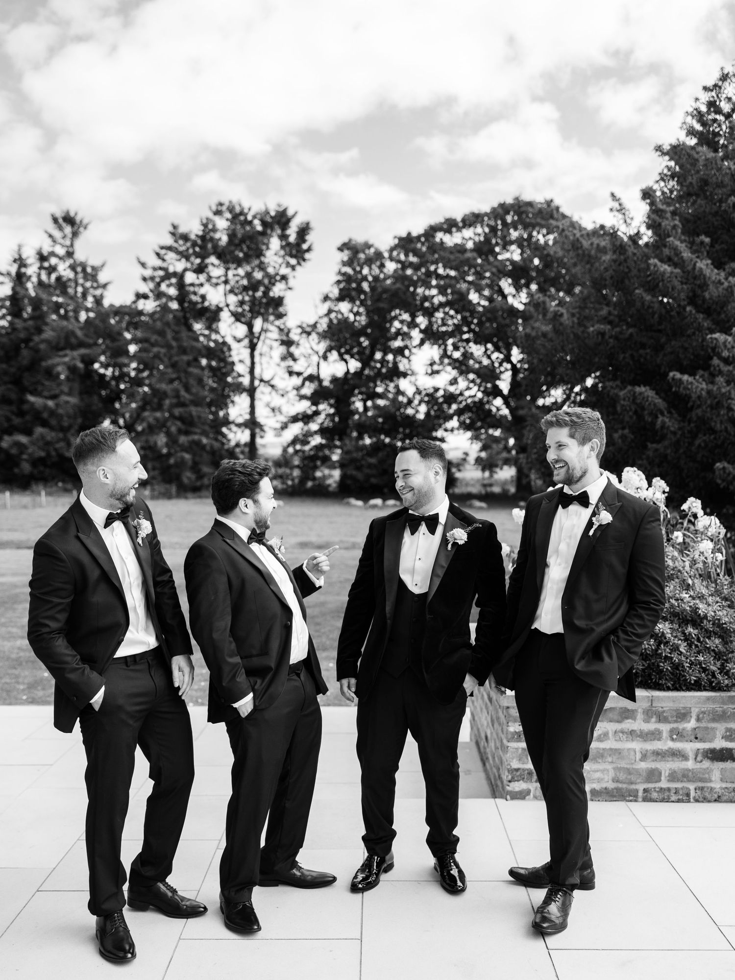 Groom and groomsmen in black tuxedos laughing together outdoors in black and white photo.