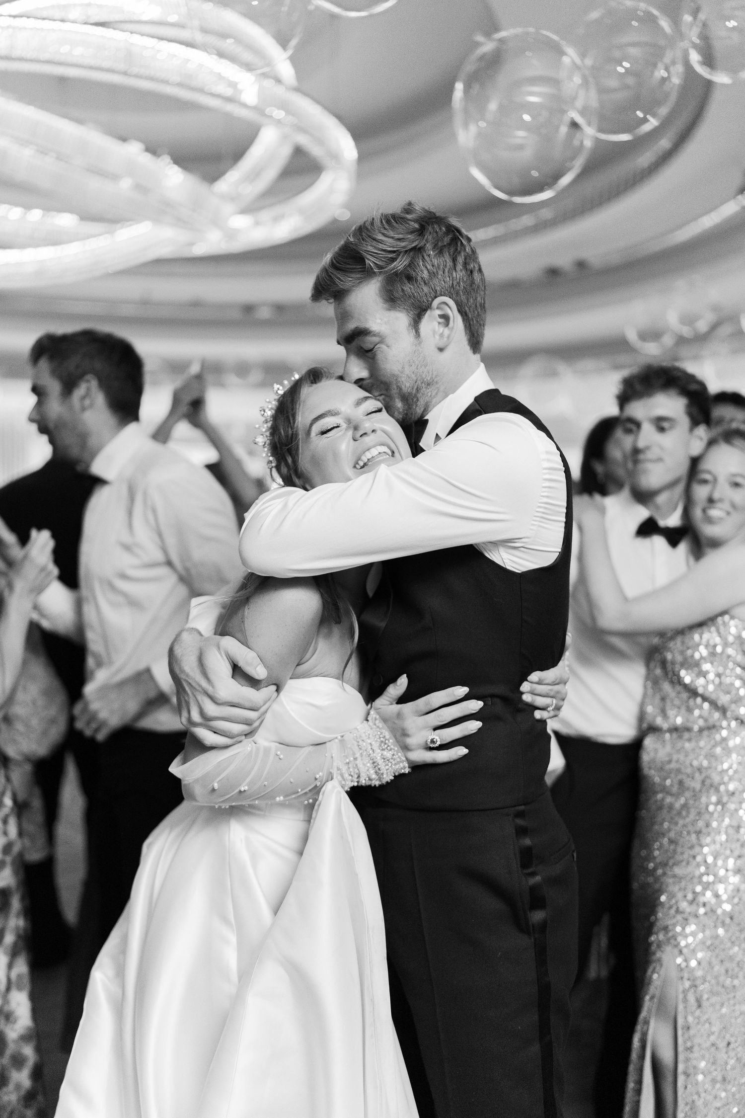 Black and white photo of bride and groom sharing first dance, embracing joyfully, with wedding guests dancing in background.