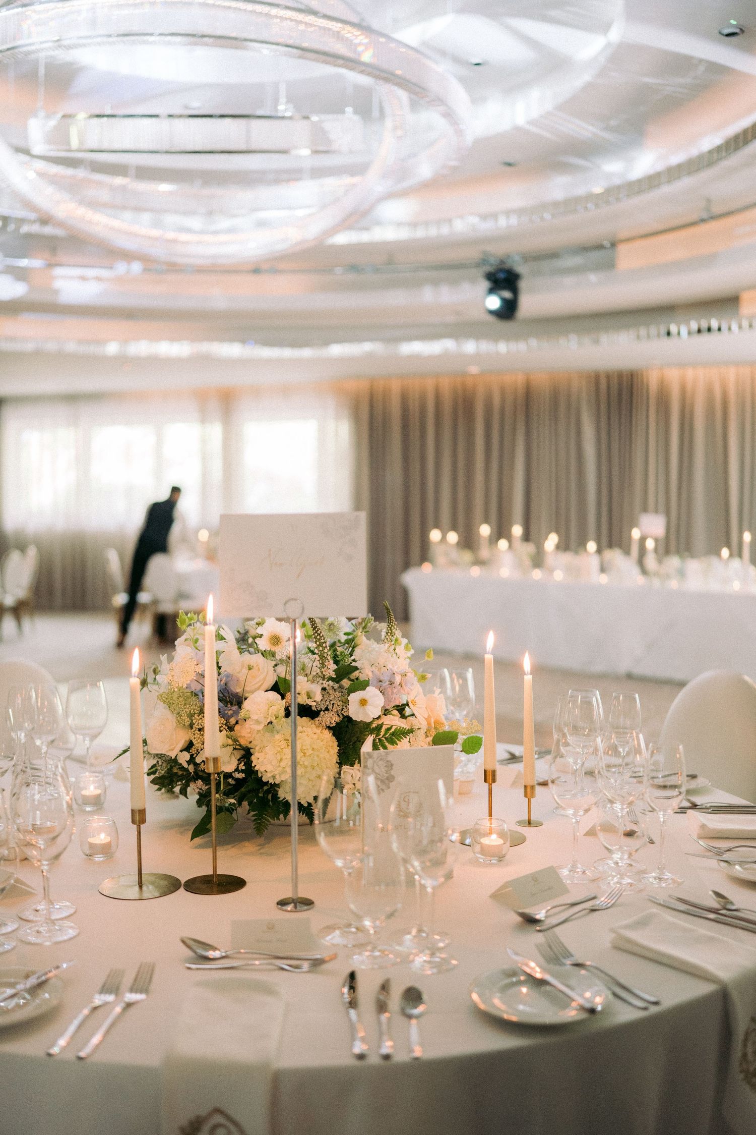 Luxurious wedding reception ballroom with candlelit round tables, floral centerpieces, and crystal ceiling lights.