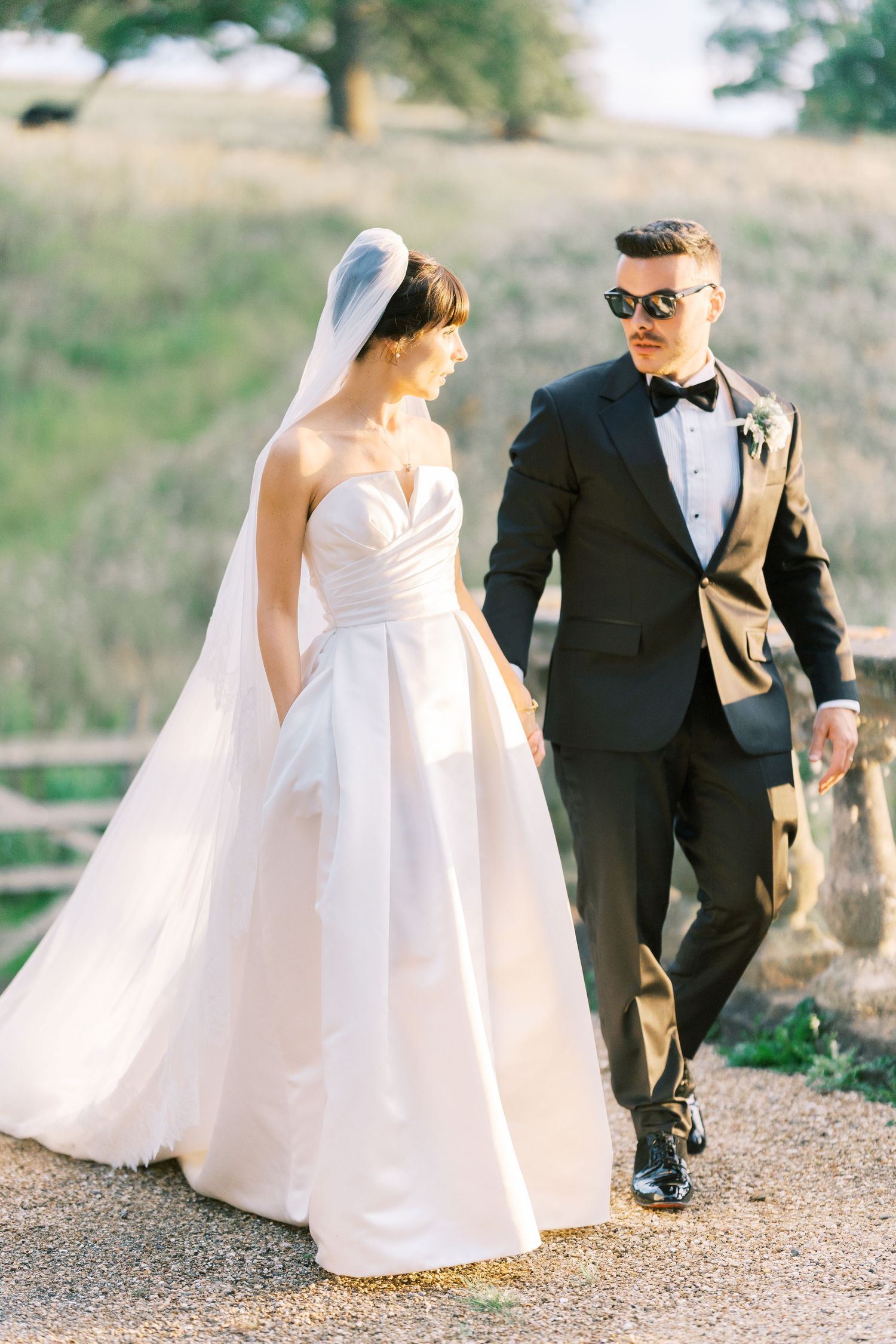 Bride in strapless ballgown and groom in black tuxedo walk hand in hand outdoors on wedding day.