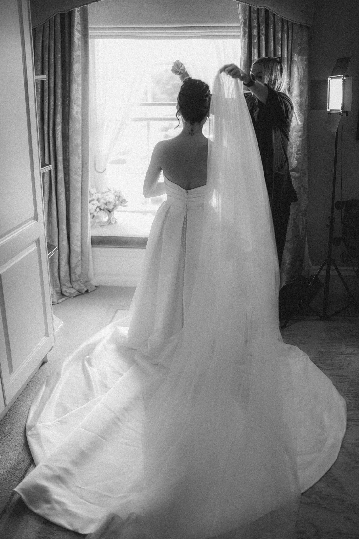 Black and white photo of bride in strapless ball gown with long veil being adjusted by stylist near window.