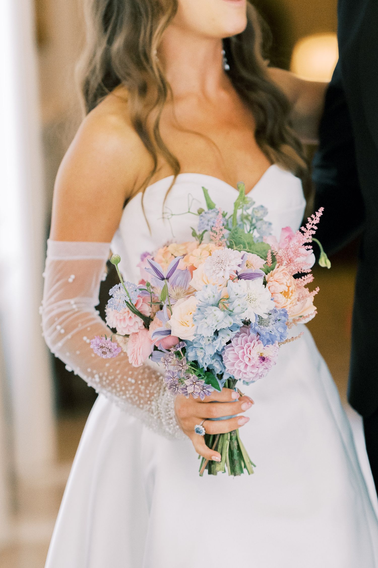 Bride in strapless white gown with sheer gloves holding a pastel bouquet of pink, blue, and purple flowers.