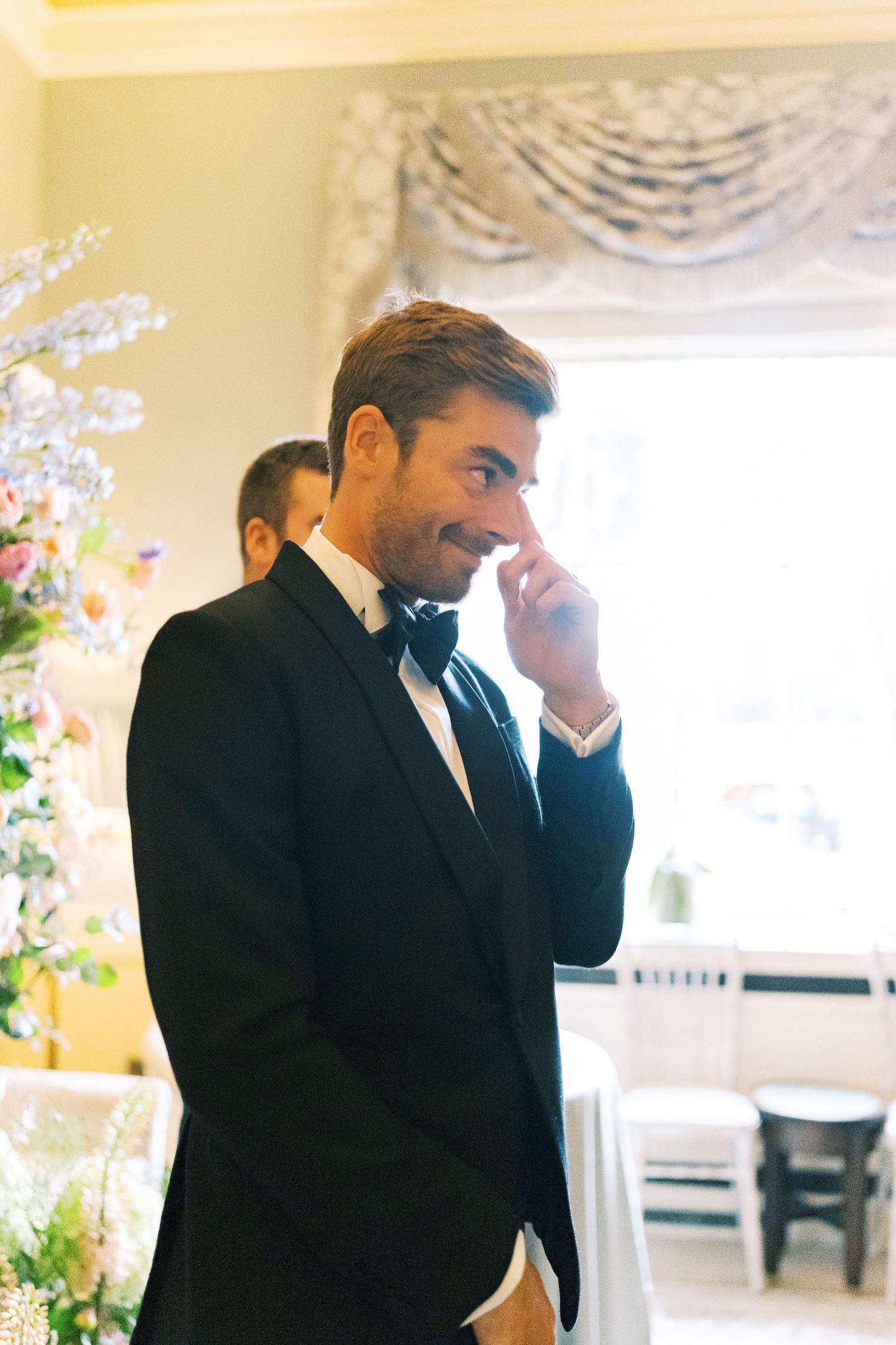 Emotional groom in black tuxedo and bow tie wiping a tear at wedding ceremony with floral arrangements.