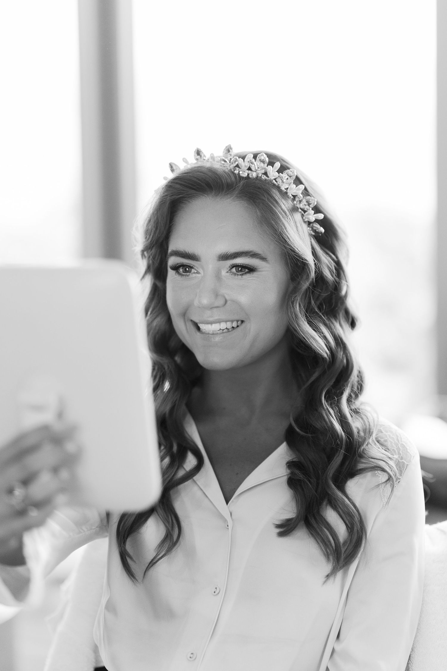 Smiling bride wearing a crystal tiara takes a selfie with a tablet, showcasing her curly hair and bridal look.