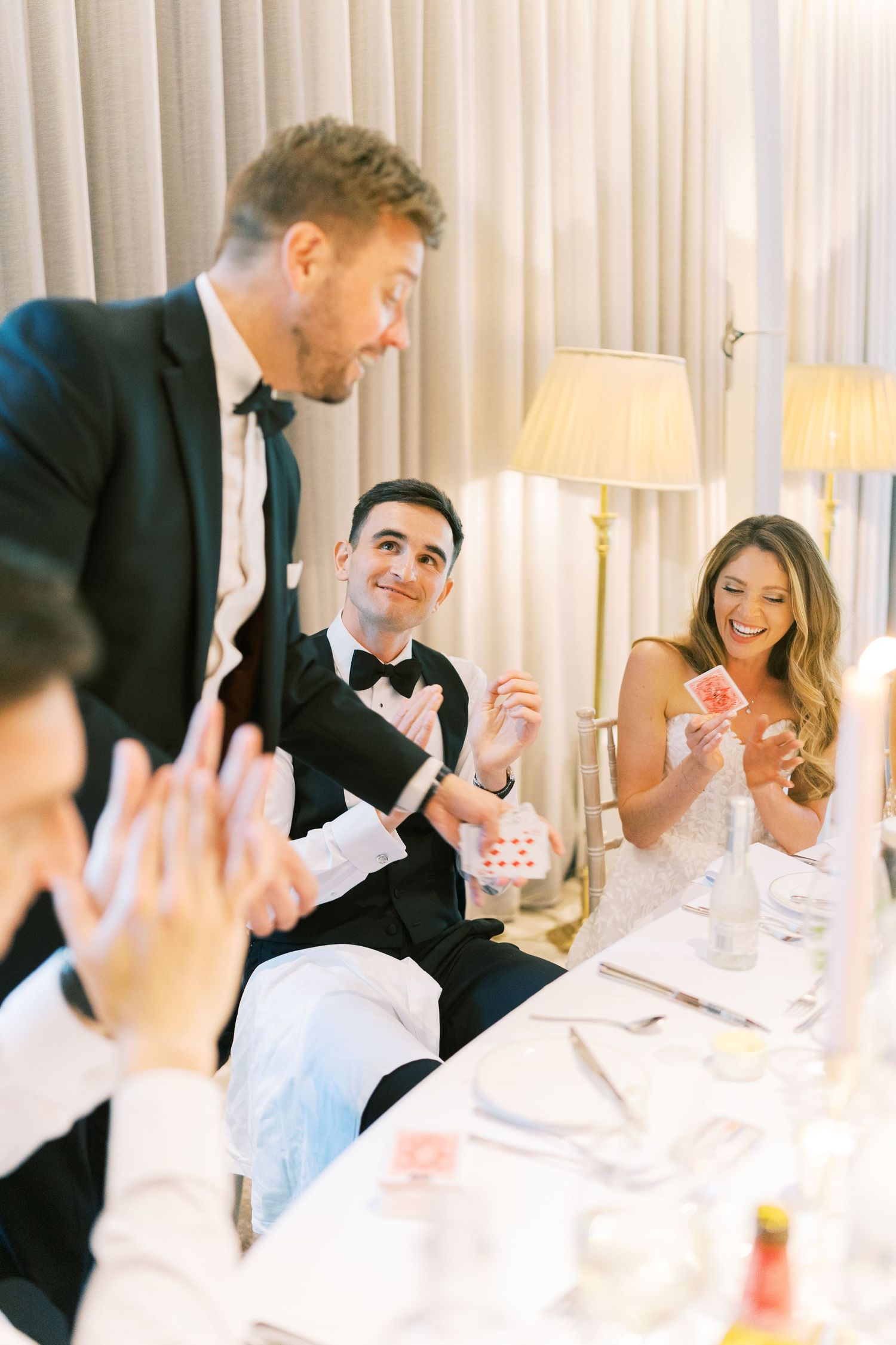 Magician performs card trick for amazed bride and groom at candlelit wedding reception table.