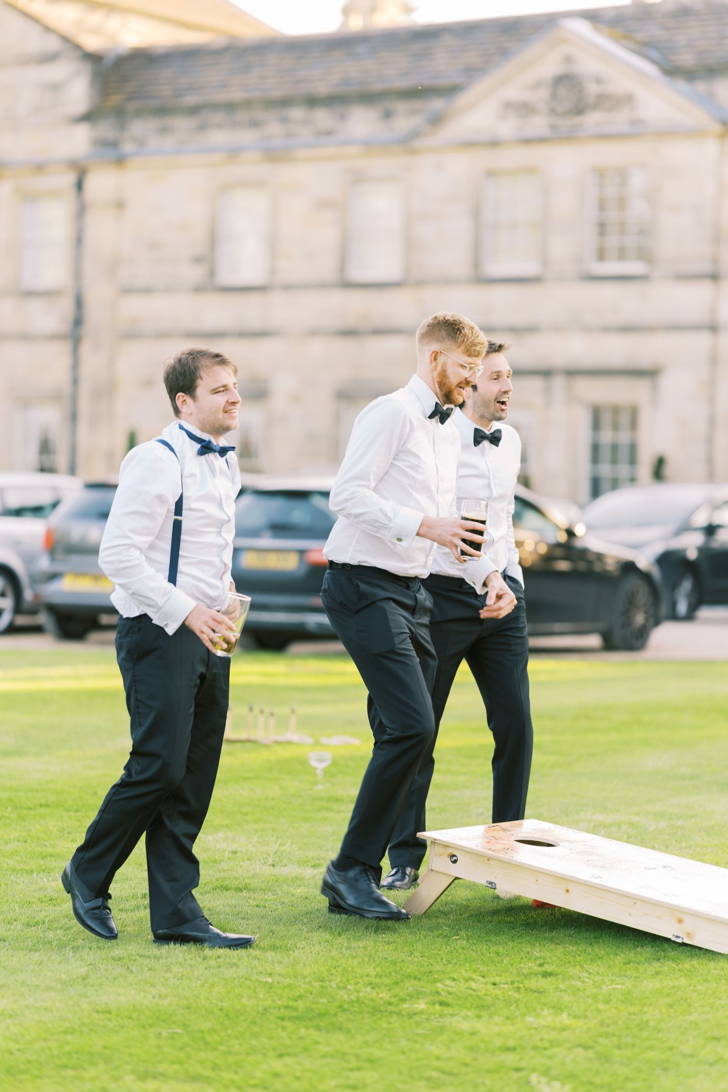 Groomsmen in white shirts and black bow ties playing cornhole on a lawn outside an elegant estate venue.
