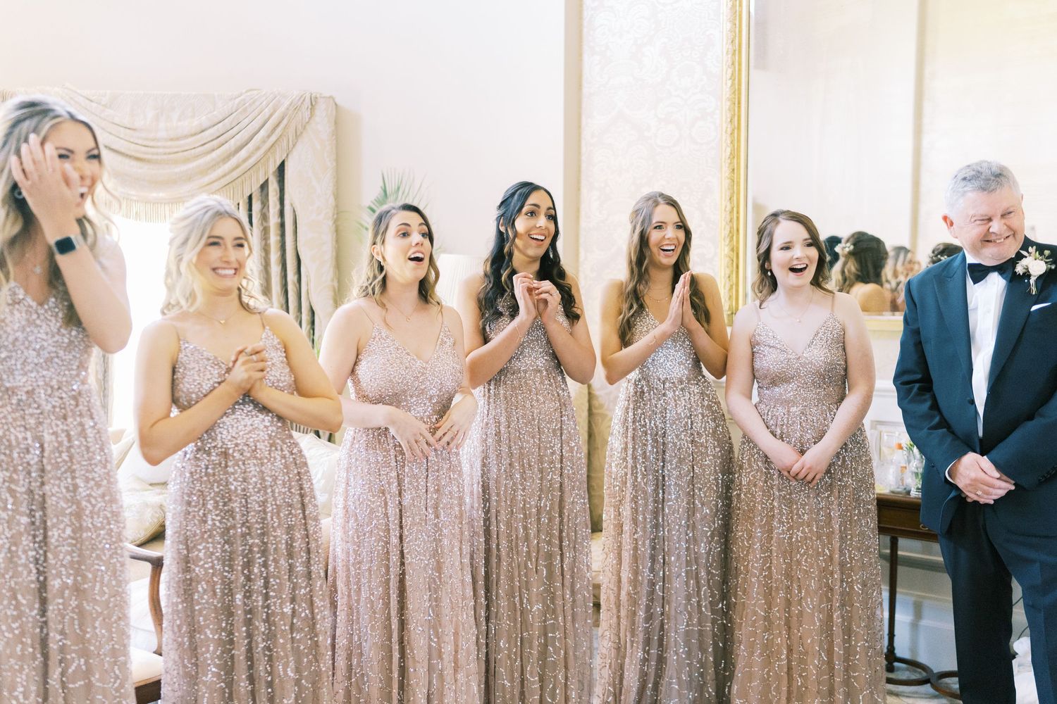 Bridesmaids in rose gold sequin gowns react with joy beside a man in a tuxedo at a wedding venue.