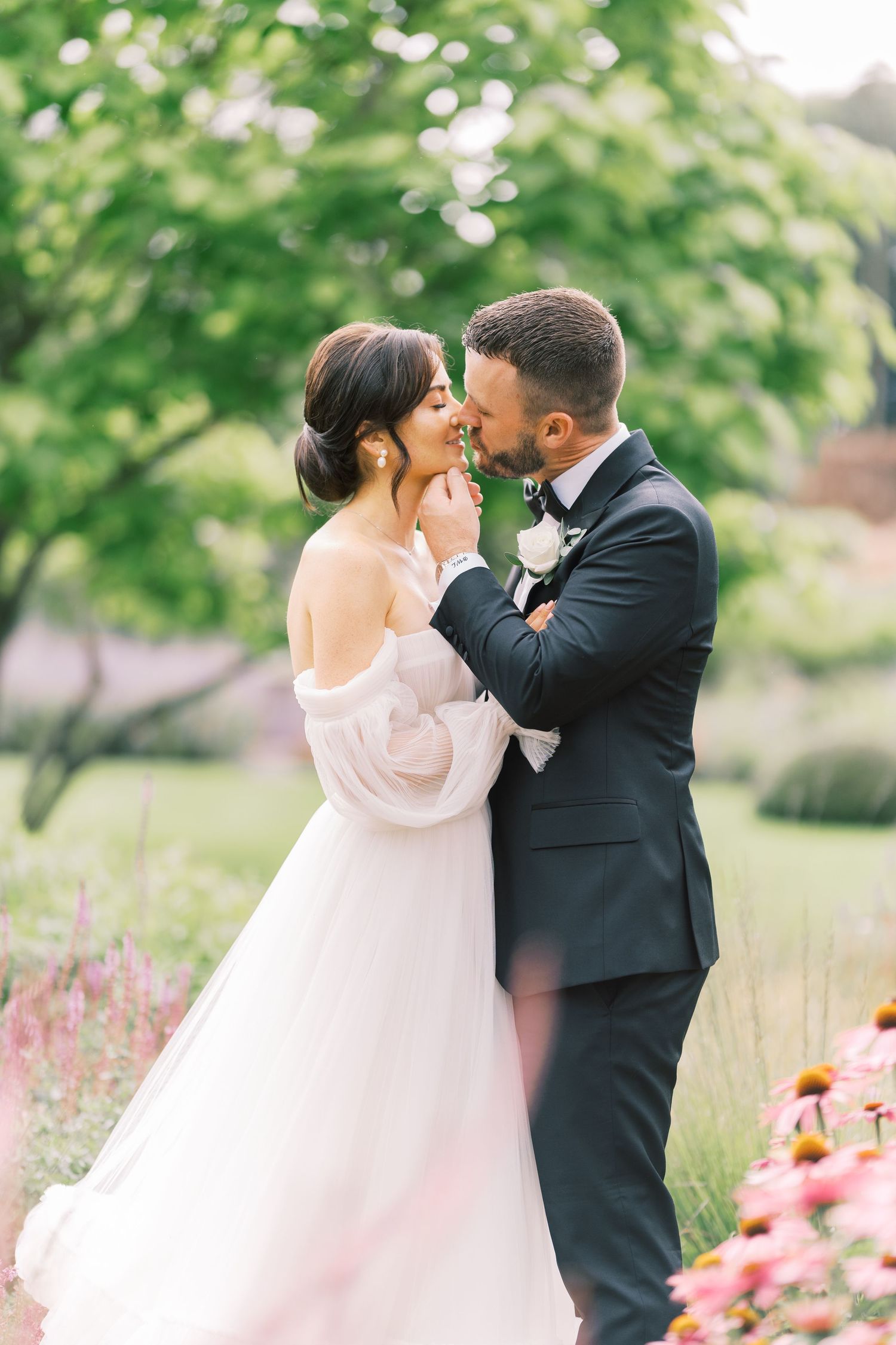 A bride and groom share a romantic kiss in a lush garden, she in an off-shoulder gown, he in a dark suit.