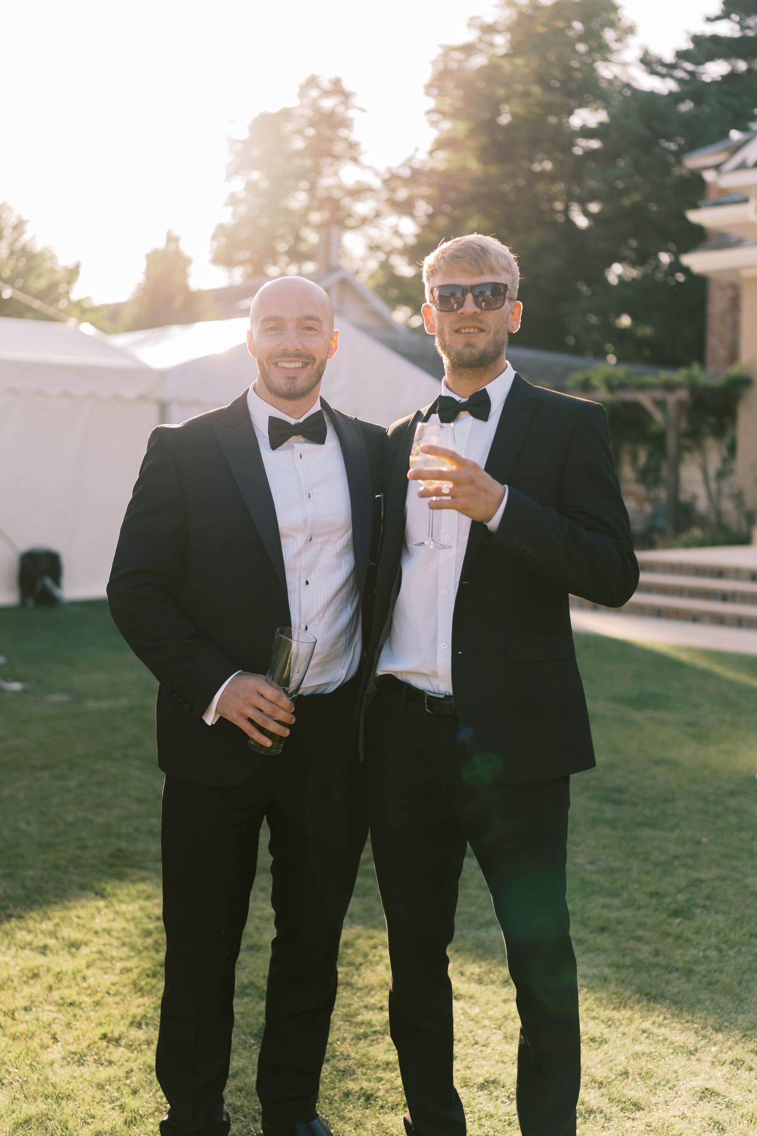 Two groomsmen in black tuxedos with bow ties holding champagne glasses on sunlit lawn at wedding venue.