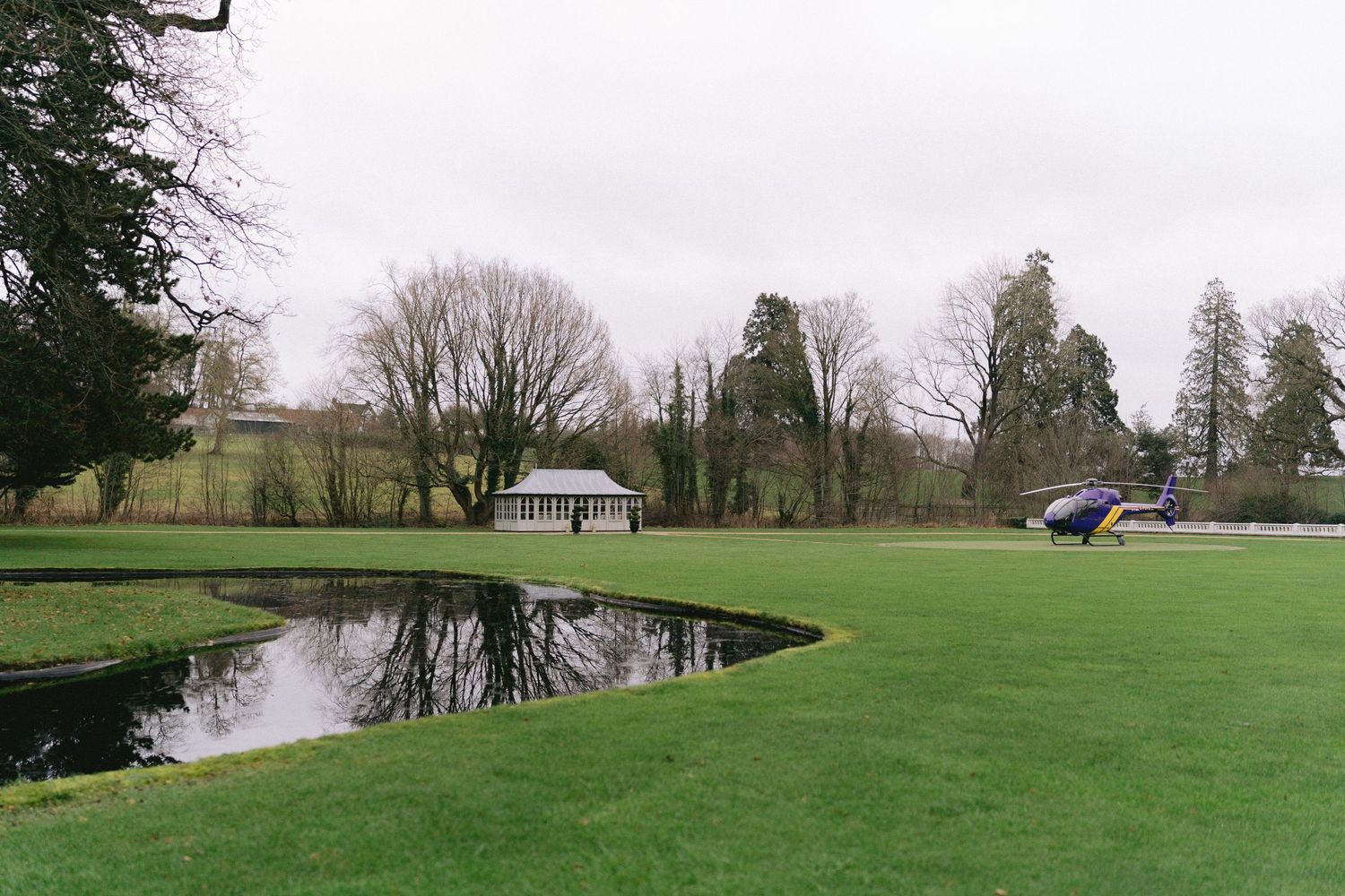 Serene English garden with a dark reflective pond, manicured lawn, and a white pavilion surrounded by bare winter trees.