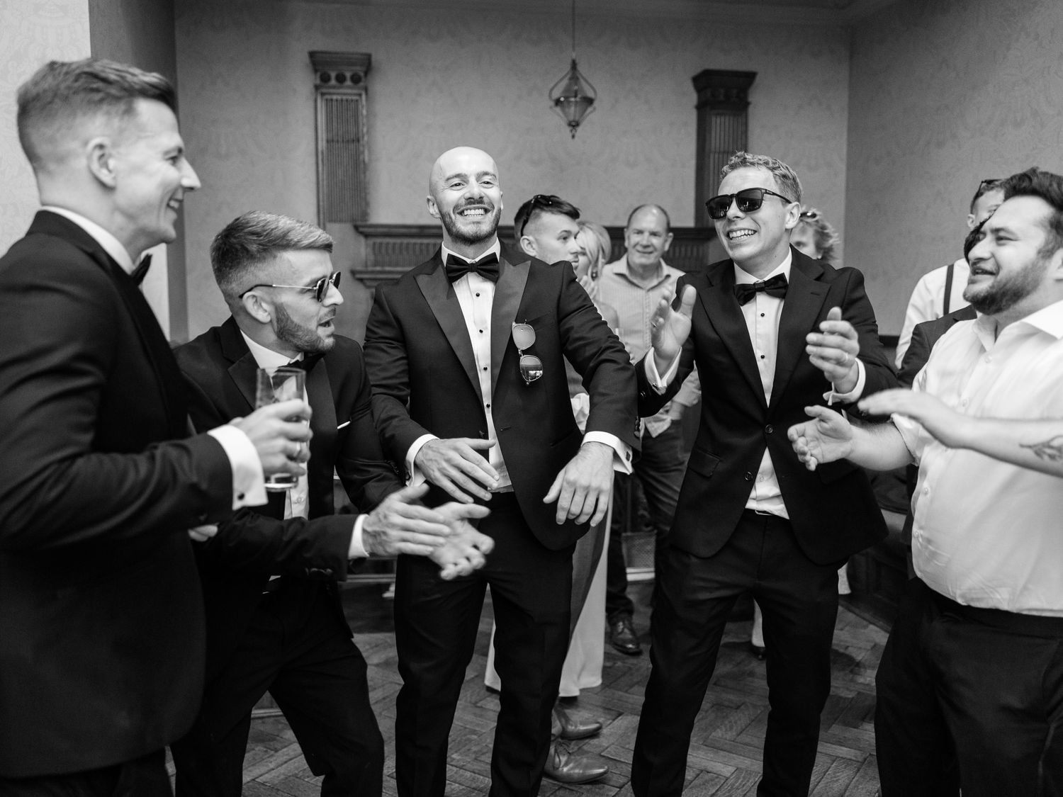 Groom celebrating with groomsmen in tuxedos, laughing and dancing at wedding reception.