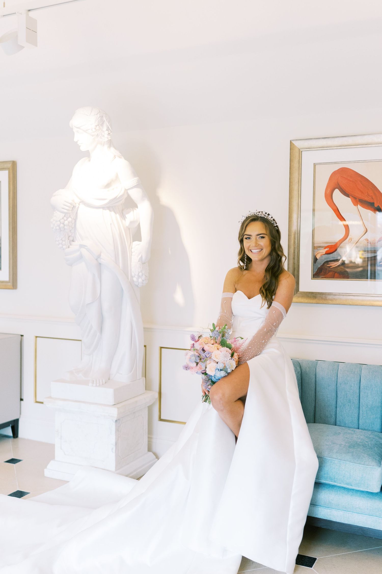 Bride in white off-shoulder gown holds pastel bouquet beside classical statue, with flamingo art and blue sofa.