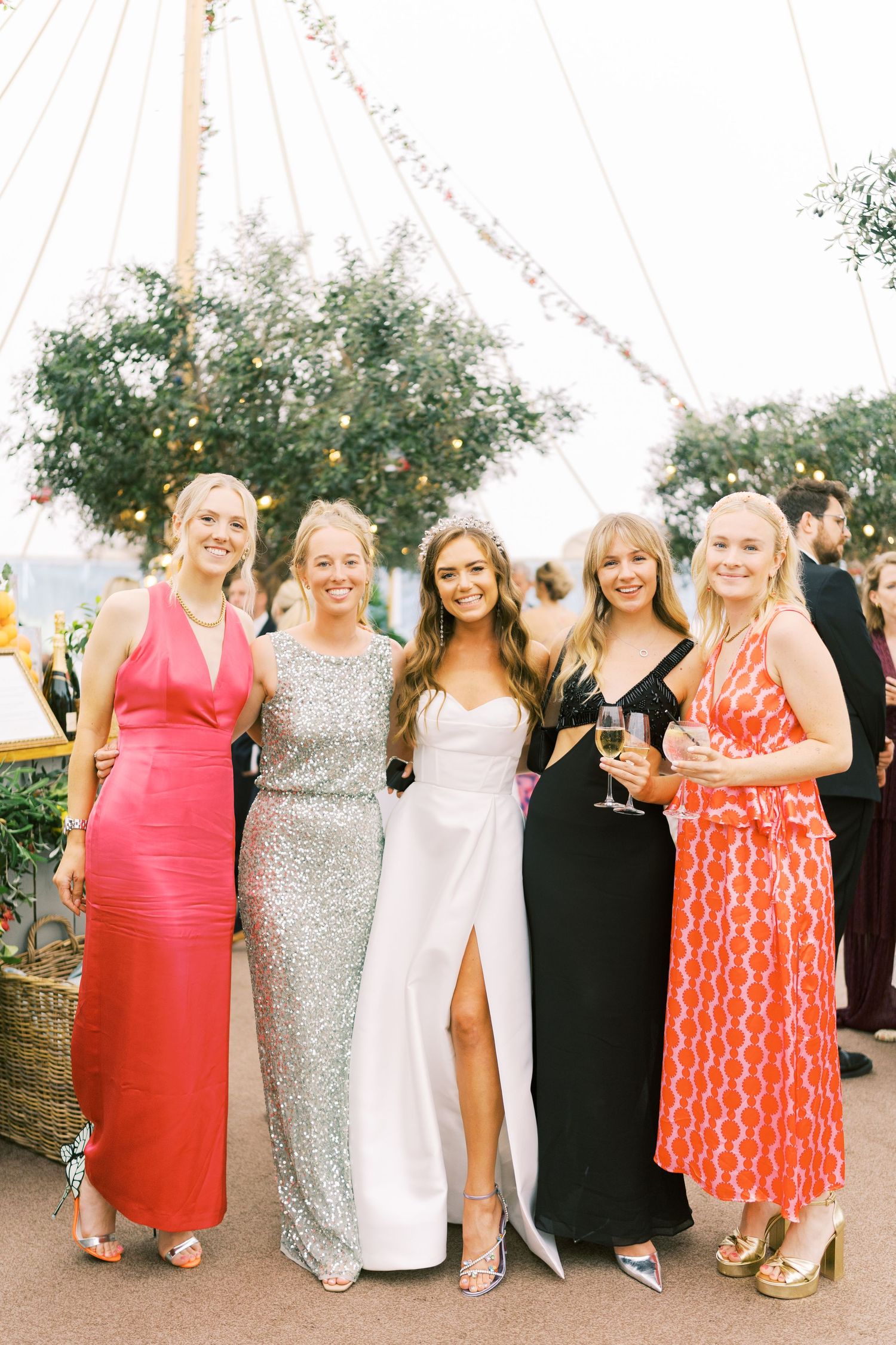 Five women in elegant formal gowns pose together smiling at an outdoor wedding reception under a decorated tent.