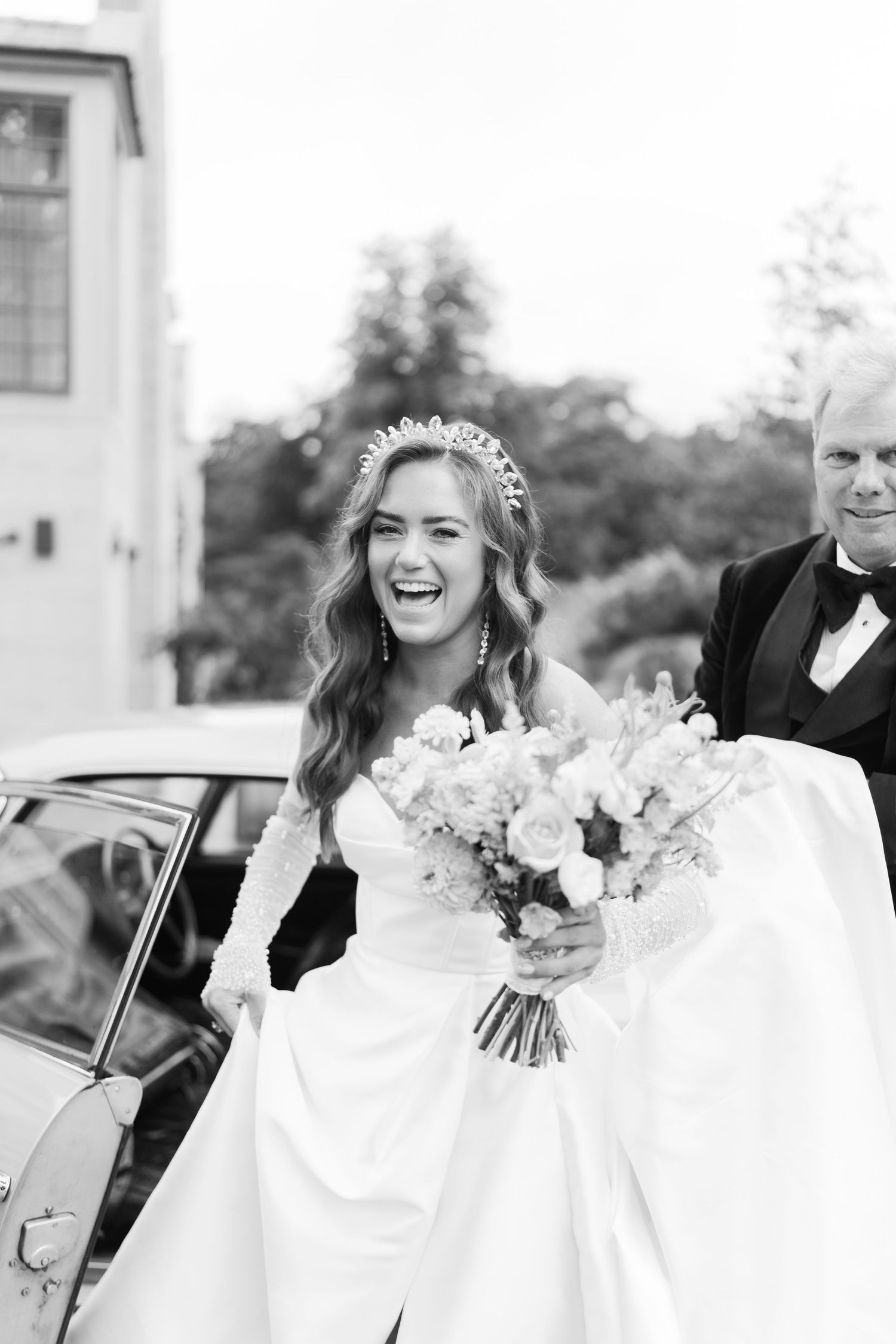 Joyful bride in tiara and white gown holding bouquet, laughing beside vintage car with older gentleman nearby.
