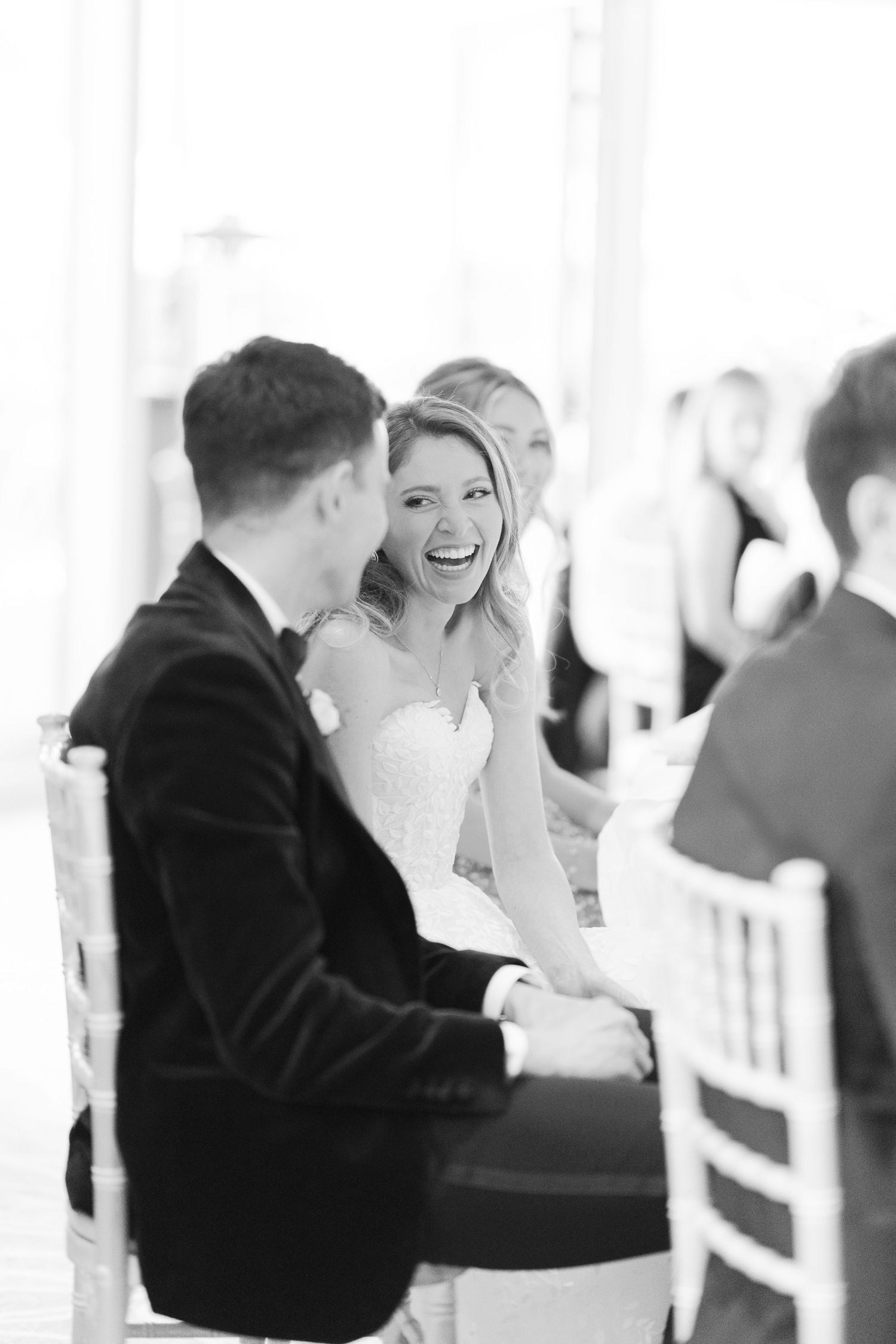 Black and white photo of a laughing bride in a strapless gown seated beside her groom during a wedding ceremony.