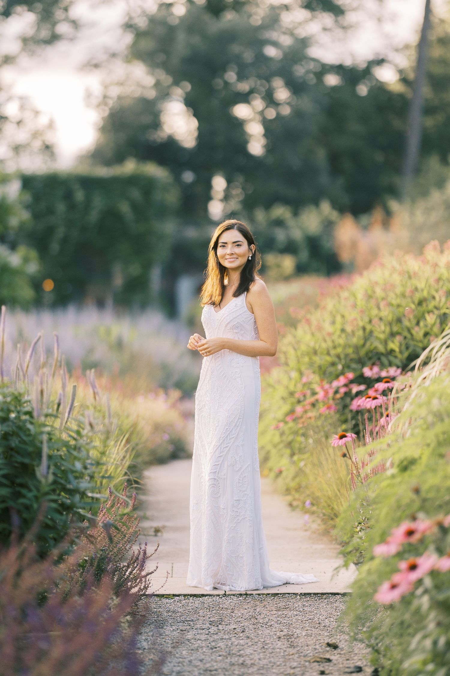 A smiling bride in a white lace gown stands on a garden path surrounded by colorful blooming flowers.