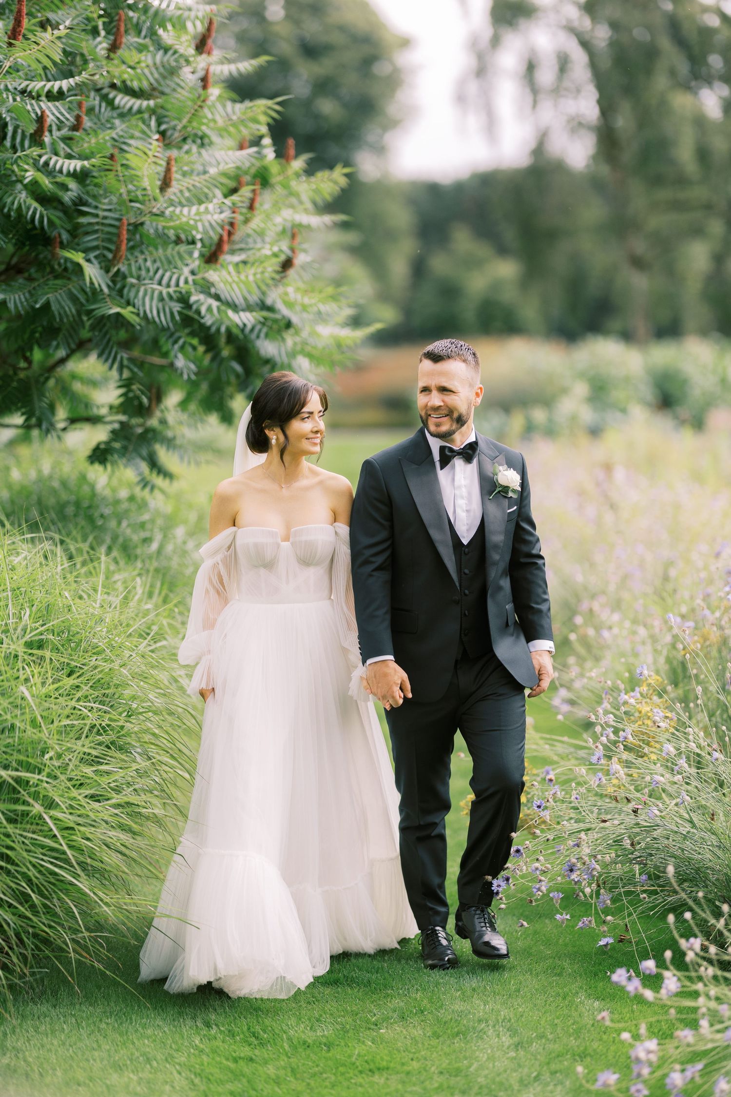 A smiling bride and groom walk hand in hand through a lush garden, she in an off-shoulder gown, he in a black tuxedo.