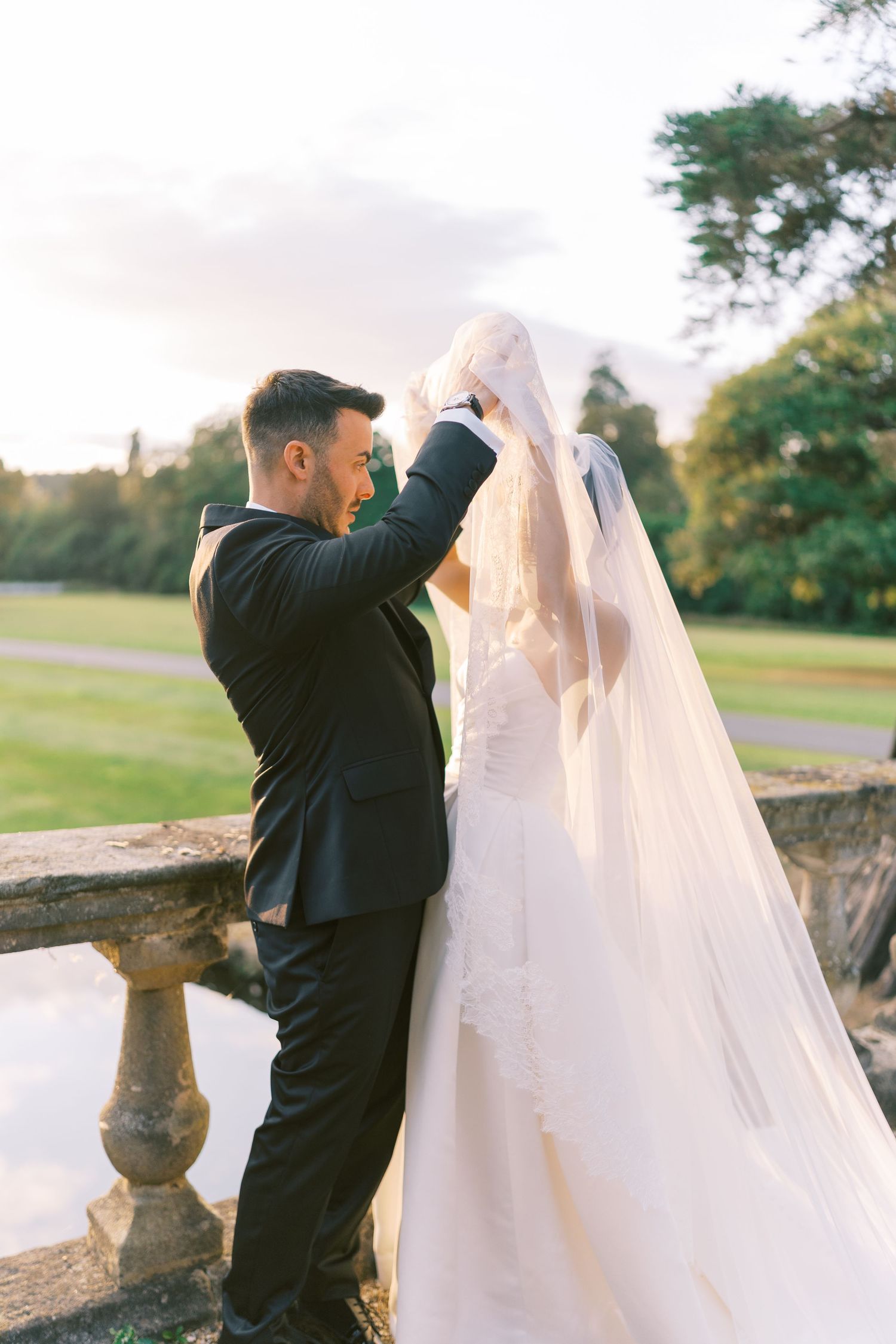 Groom in black suit adjusting brides cathedral veil on stone terrace at sunset wedding.